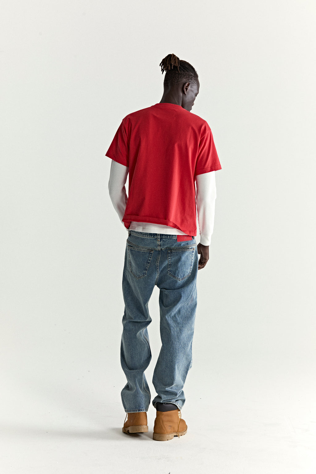 Man in red ARAIGNÉE TEE over white long-sleeve, blue jeans, and tan boots.