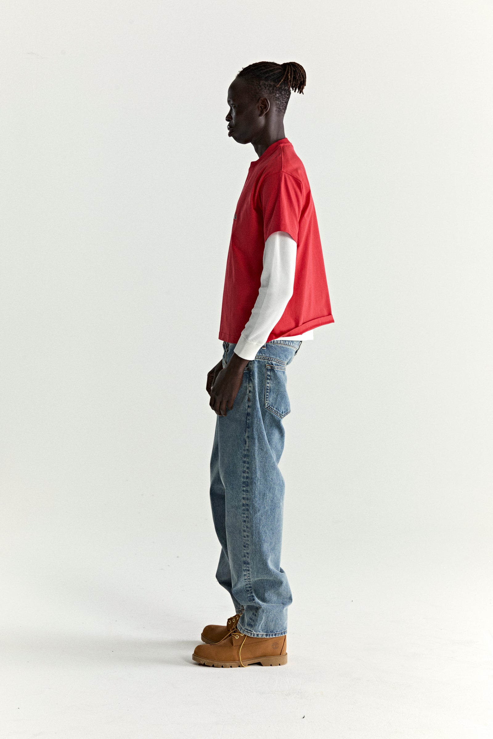 Man in red cotton ARAIGNÉE TEE with embroidered graphic, layered over a white long-sleeve, blue jeans, and brown boots.