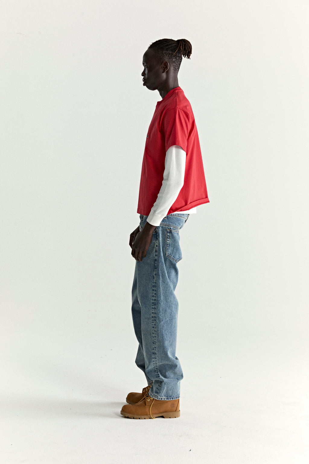 Man in red cotton ARAIGNÉE TEE with embroidered graphic, layered over a white long-sleeve, blue jeans, and brown boots.