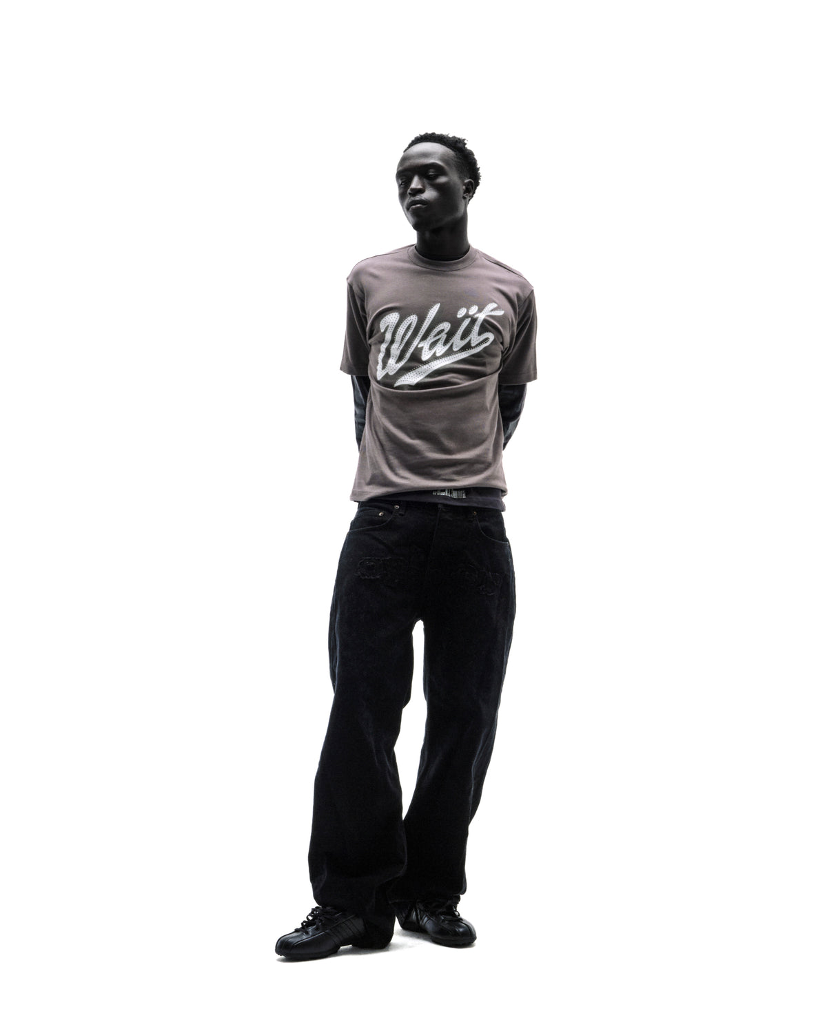 A model wearing a grey graphic t-shirt with the word Wait, black baggy jeans, and black sneakers against a white background.