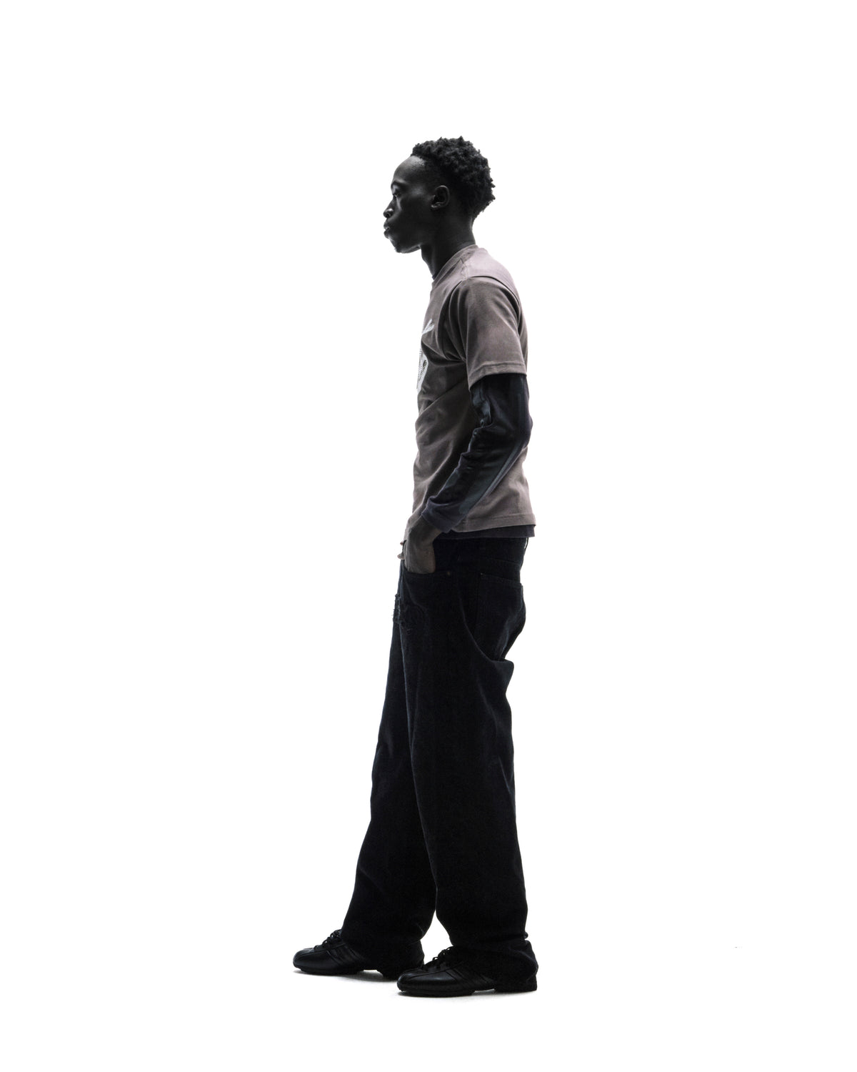 A model in profile wearing a layered grey t-shirt, black baggy jeans, and sneakers against a white background.