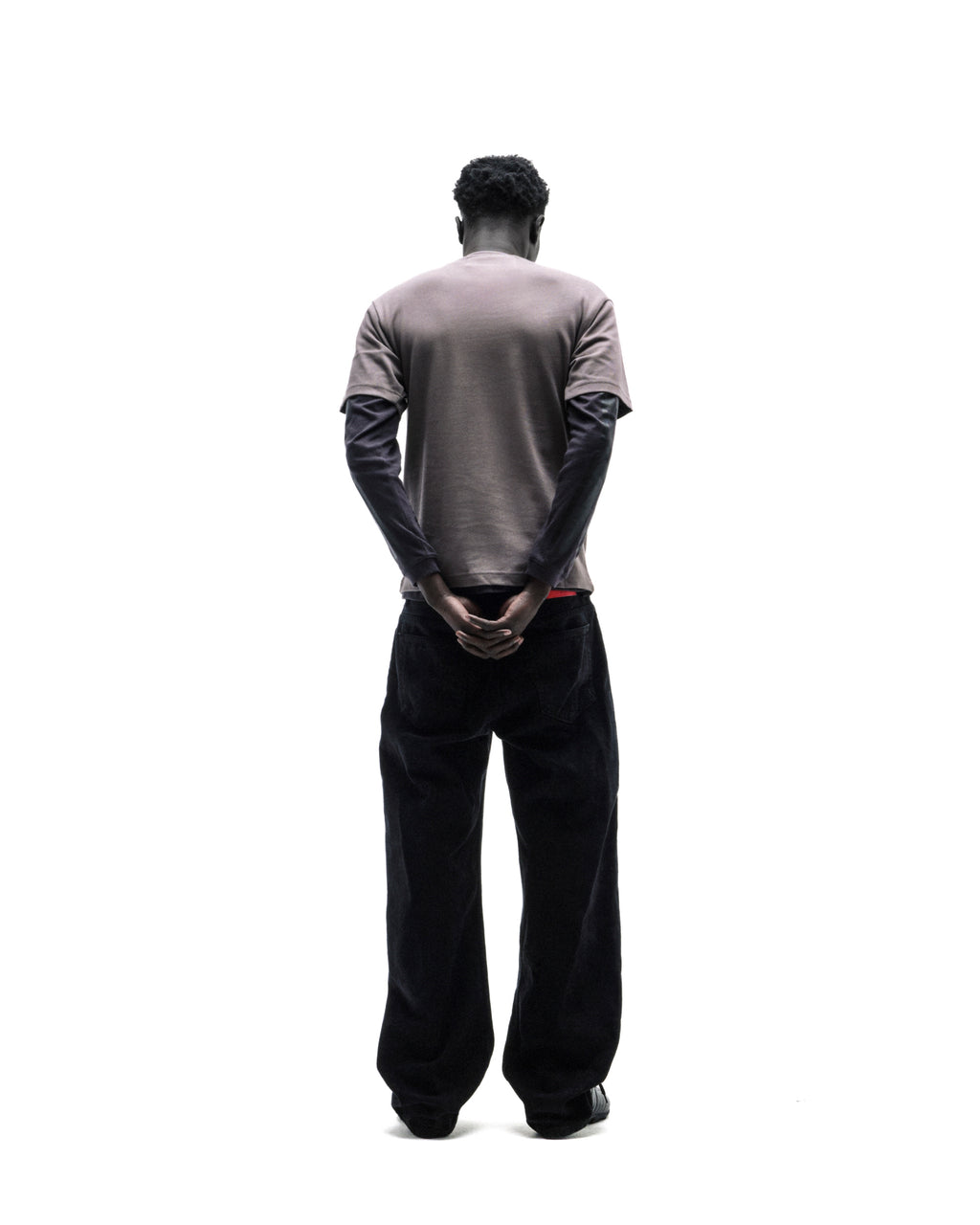 A model standing with back to camera, wearing a brown t-shirt over a black long-sleeve shirt and loose black jeans.