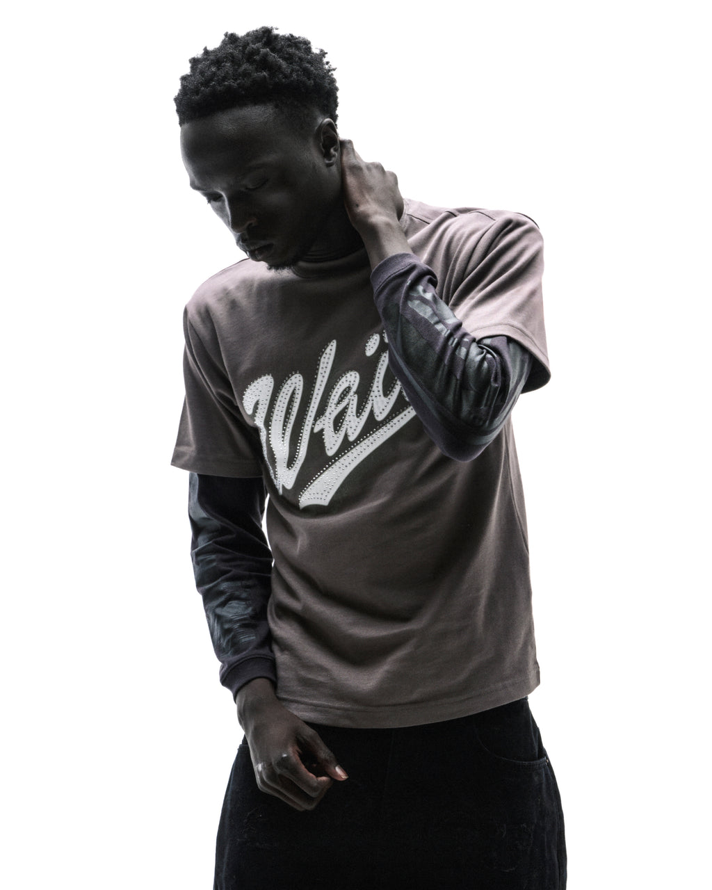 A model wearing a brown graphic t-shirt layered over a black long-sleeve shirt with a white logo against a white background.