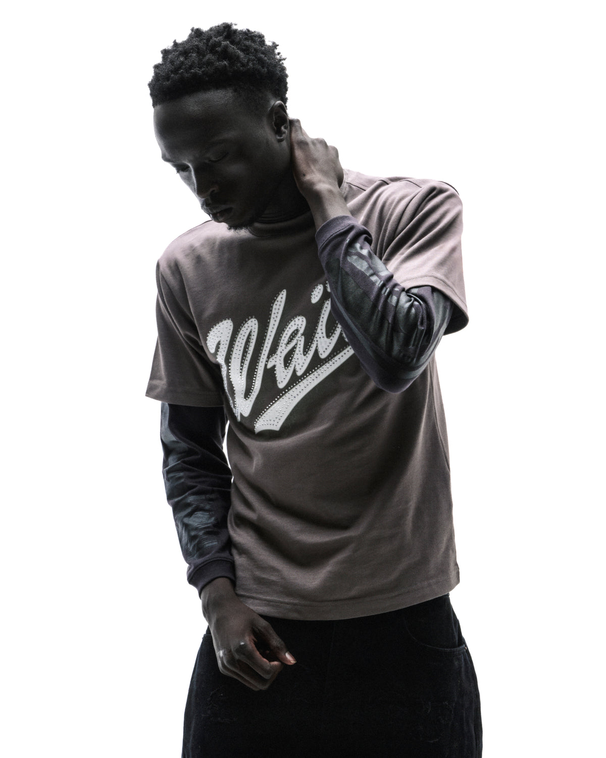A model wearing a brown graphic t-shirt layered over a black long-sleeve shirt with a white logo against a white background.