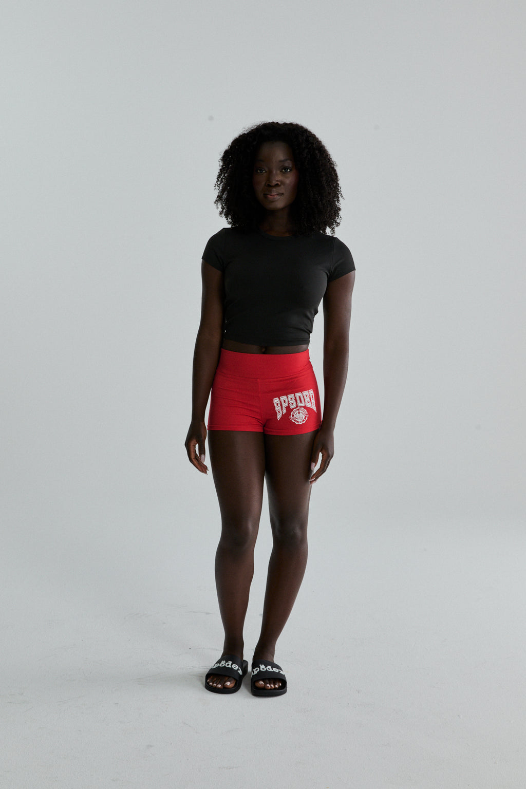 Woman wearing red stretch cotton jersey SP5DER Tiny Short with collegiate graphic and black crop top.