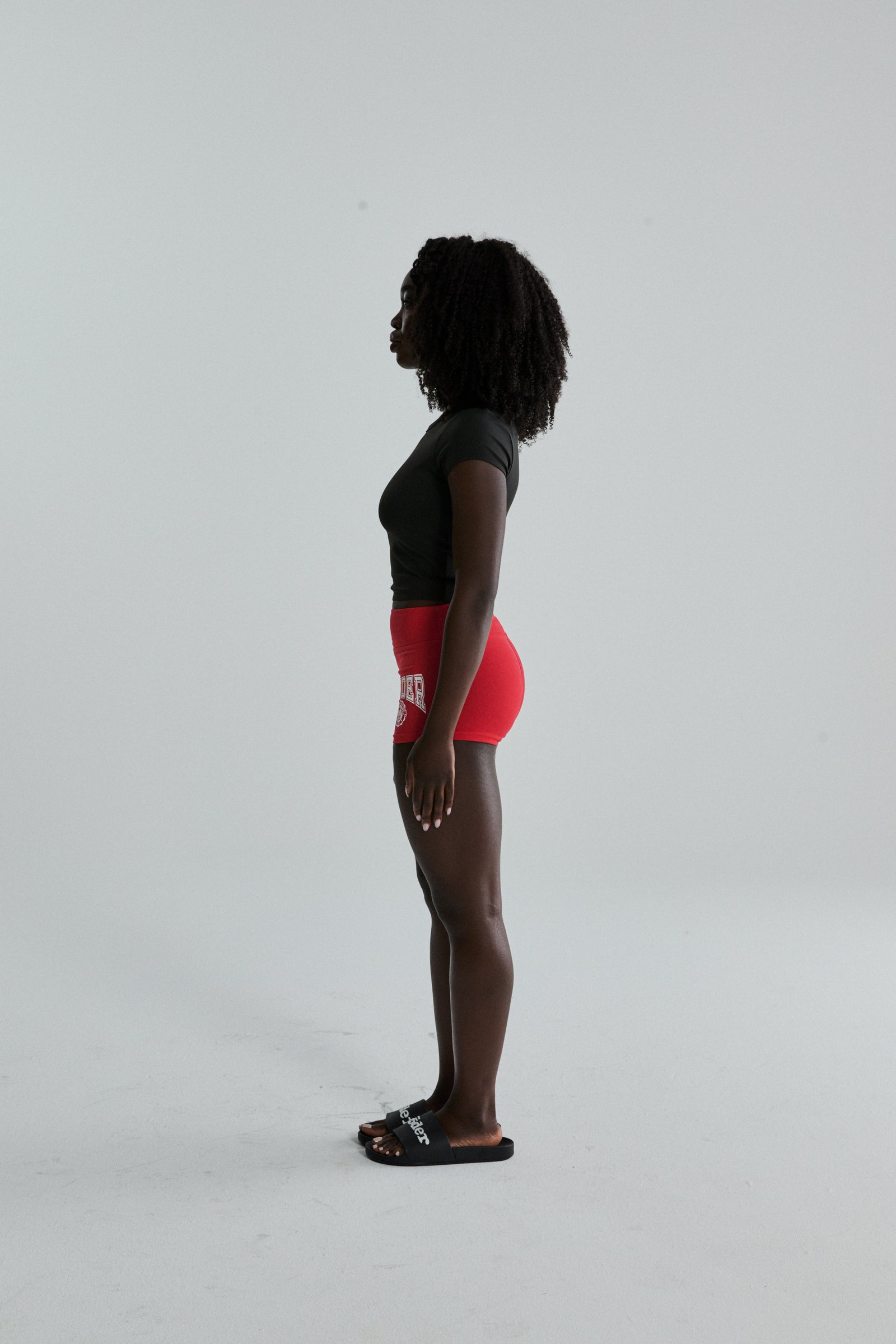 Woman in red stretch cotton jersey SP5DER Tiny Short with collegiate graphic, side profile.