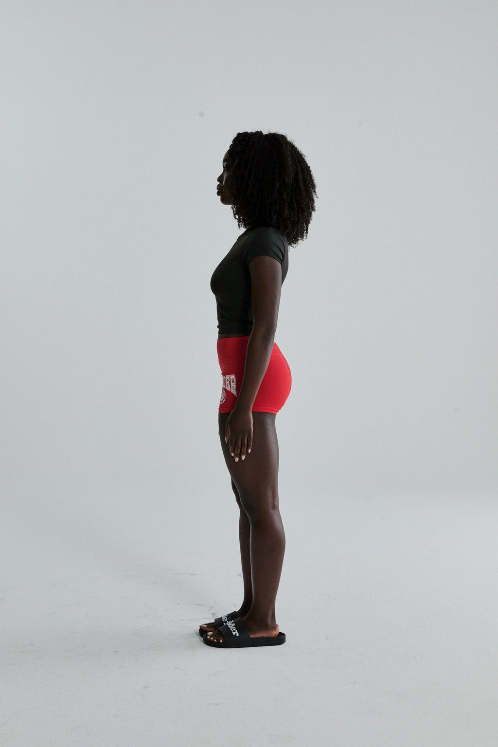 Woman in red stretch cotton jersey SP5DER Tiny Short with collegiate graphic, side profile.