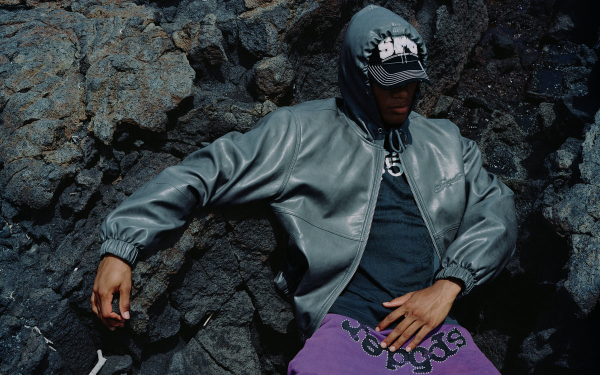 Model in a grey leather hooded jacket, black cap, and purple embellished shorts posing against dark volcanic rock.