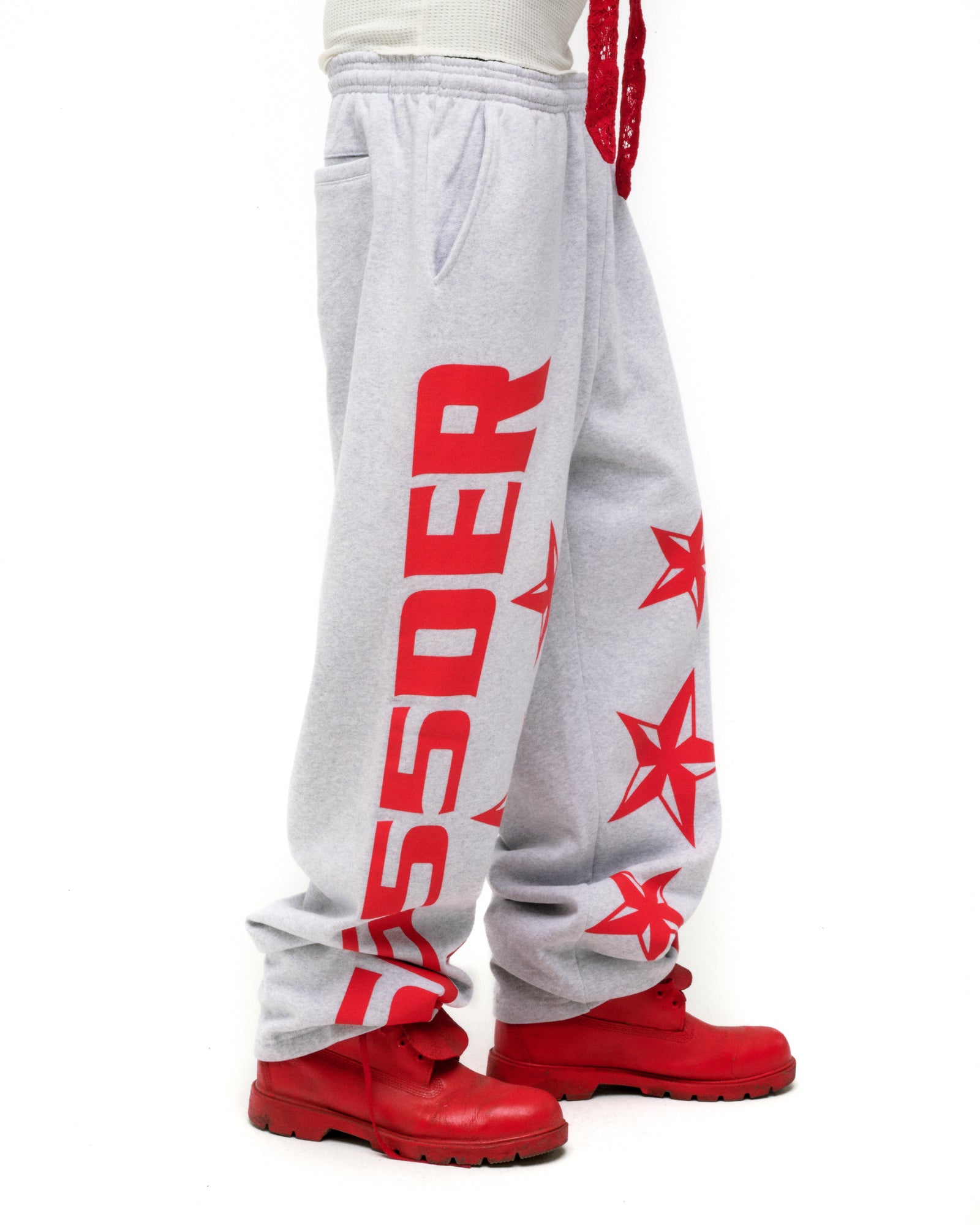 Light gray MX555 sweatpants with bold red SP5DER text and star print, wide-leg, worn with red boots.
