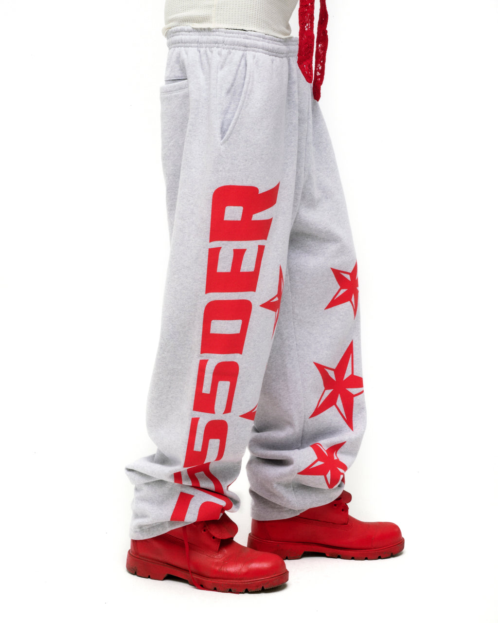 Light gray MX555 sweatpants with bold red SP5DER text and star print, wide-leg, worn with red boots.