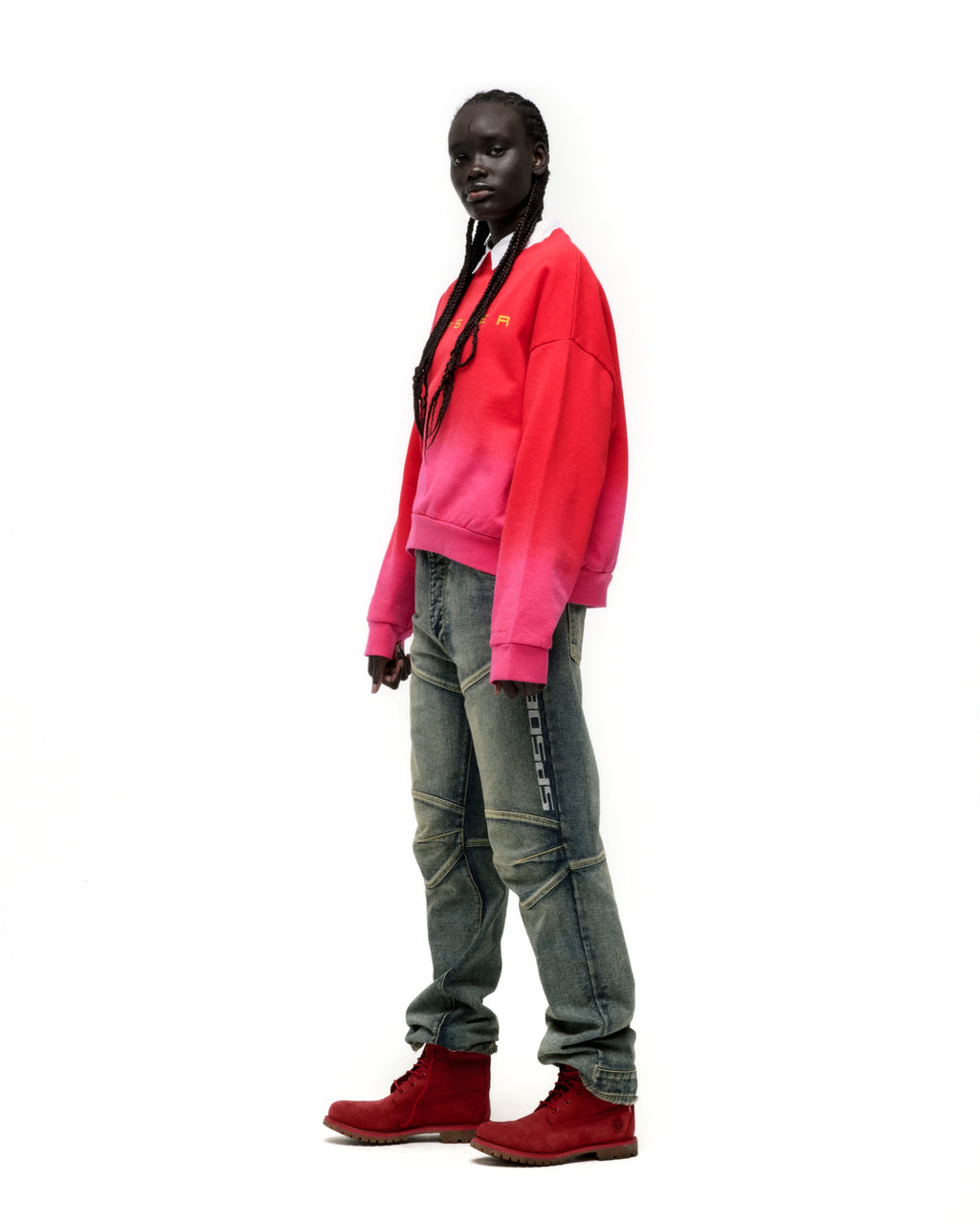 Model wearing a red gradient crewneck with SP5DER text, blue jeans, and red boots.