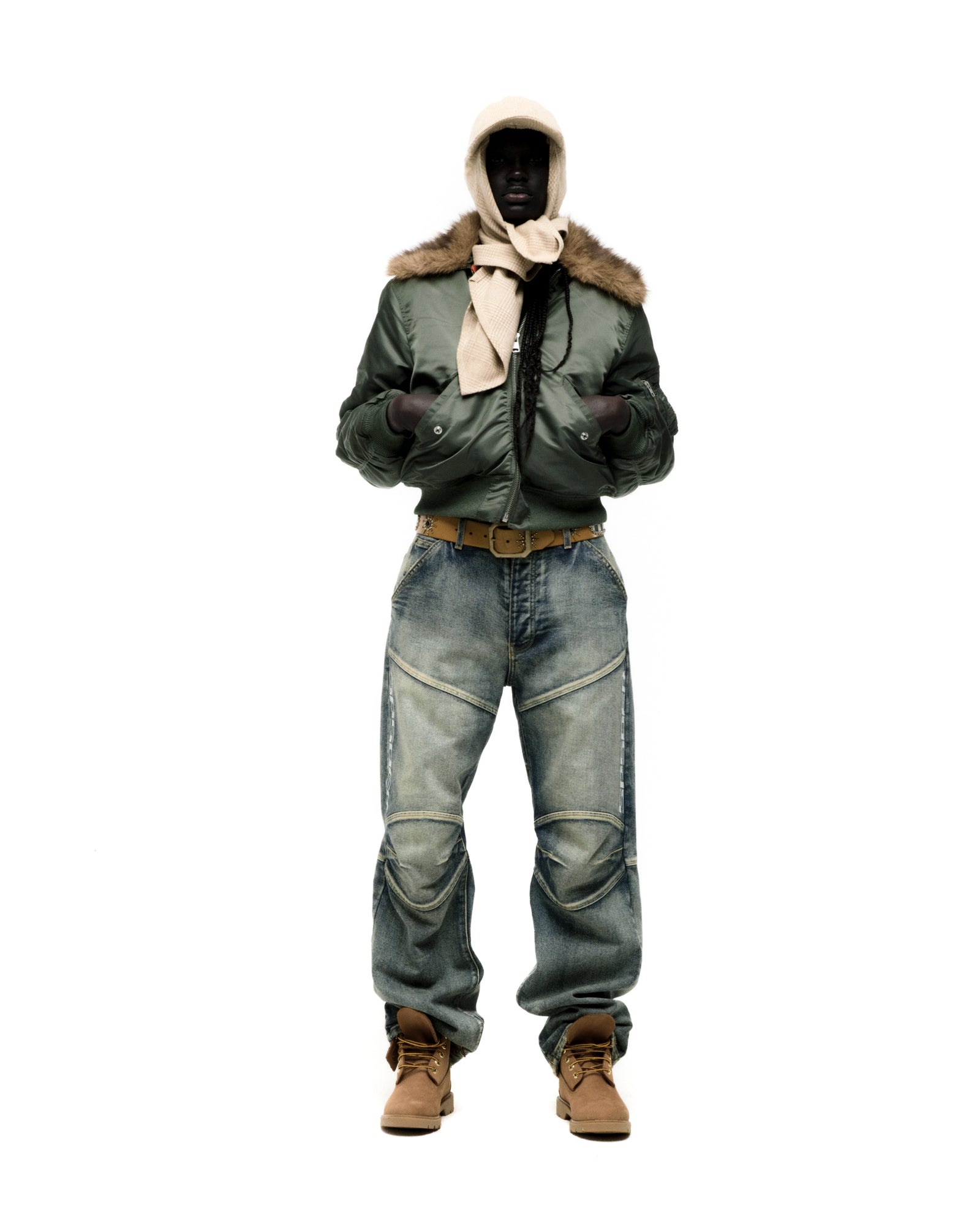 Man in olive green nylon bomber jacket with faux fur collar, blue jeans, and tan boots.