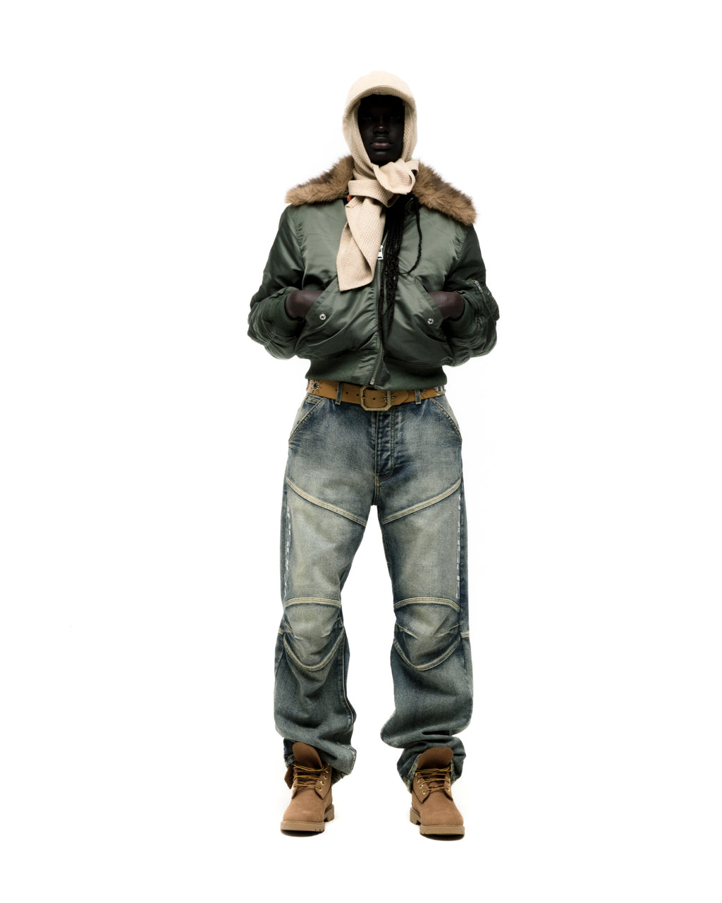 Man in olive green nylon bomber jacket with faux fur collar, blue jeans, and tan boots.