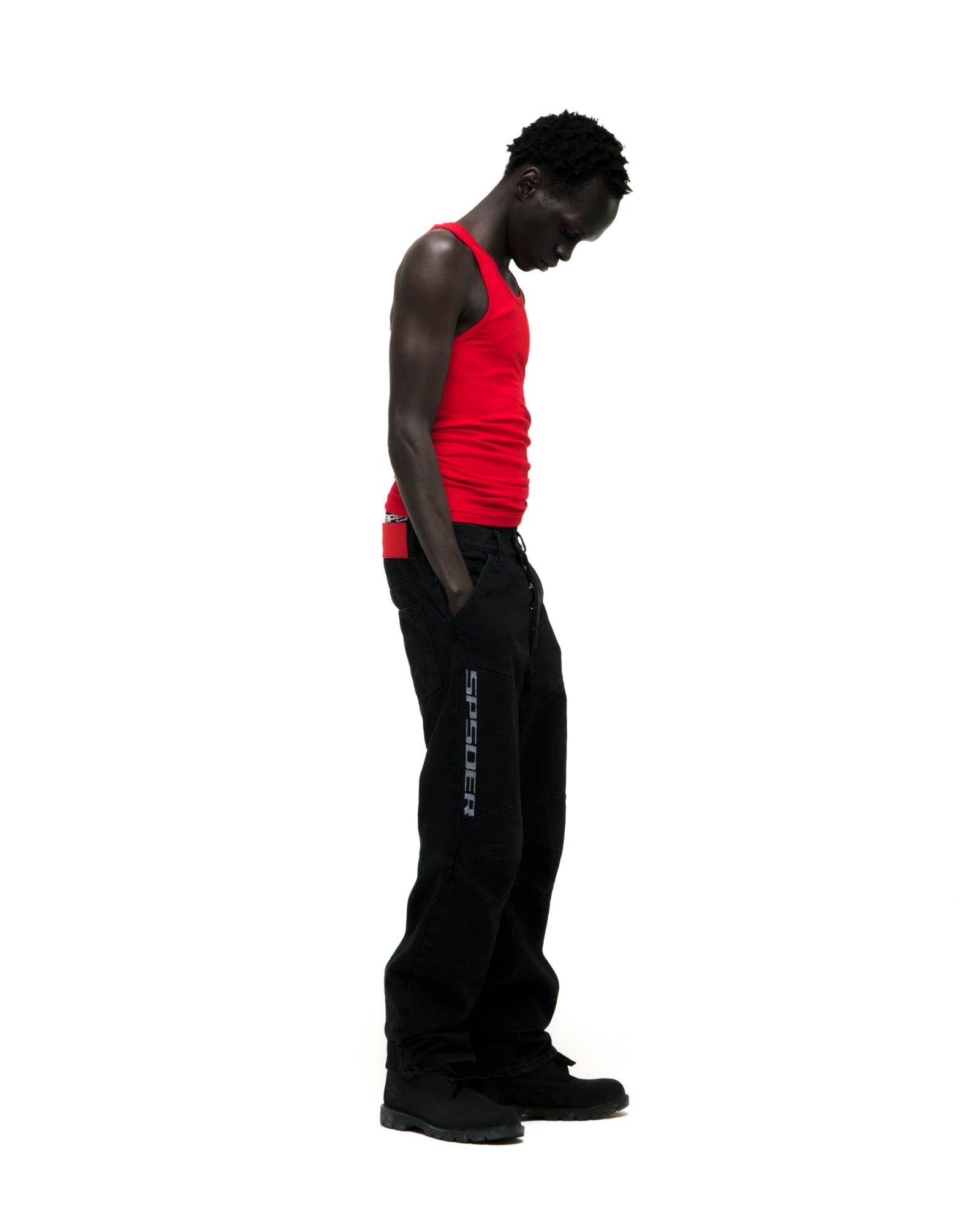 Man in red tank top and black "SP5DER" logo denim pants with paneled knees and distressed details.