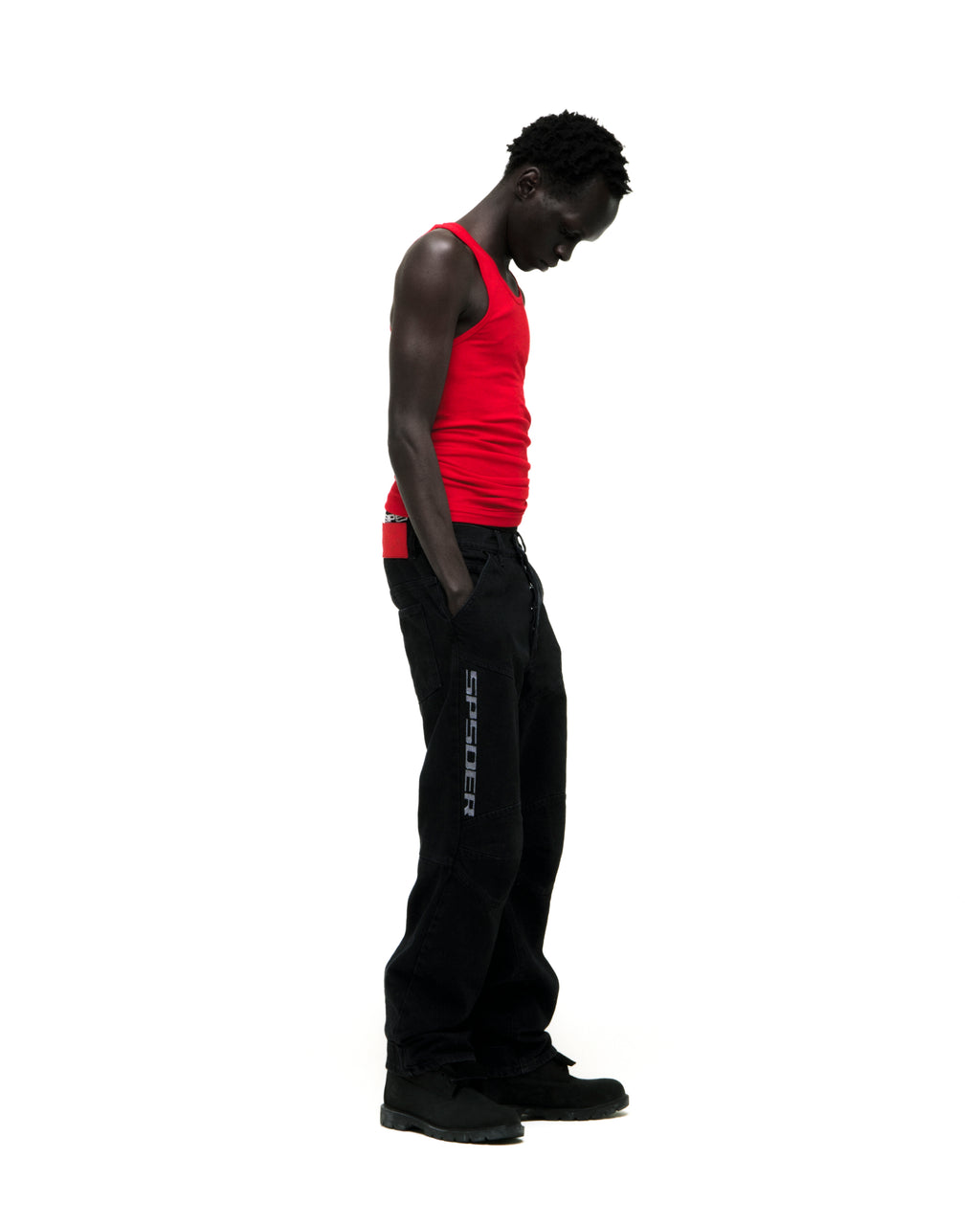 Man in red tank top and black "SP5DER" logo denim pants with paneled knees and distressed details.