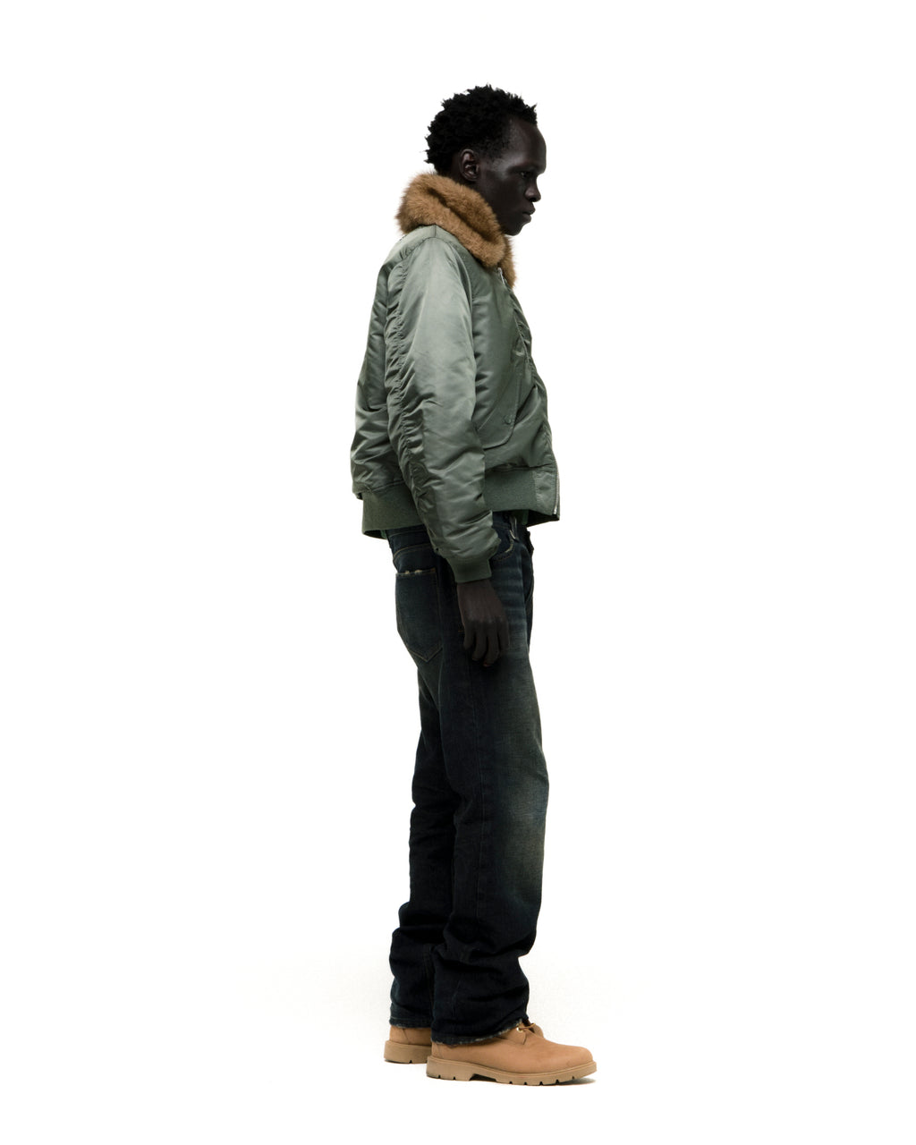 Man in olive nylon bomber jacket with faux fur collar, dark jeans, and tan boots.