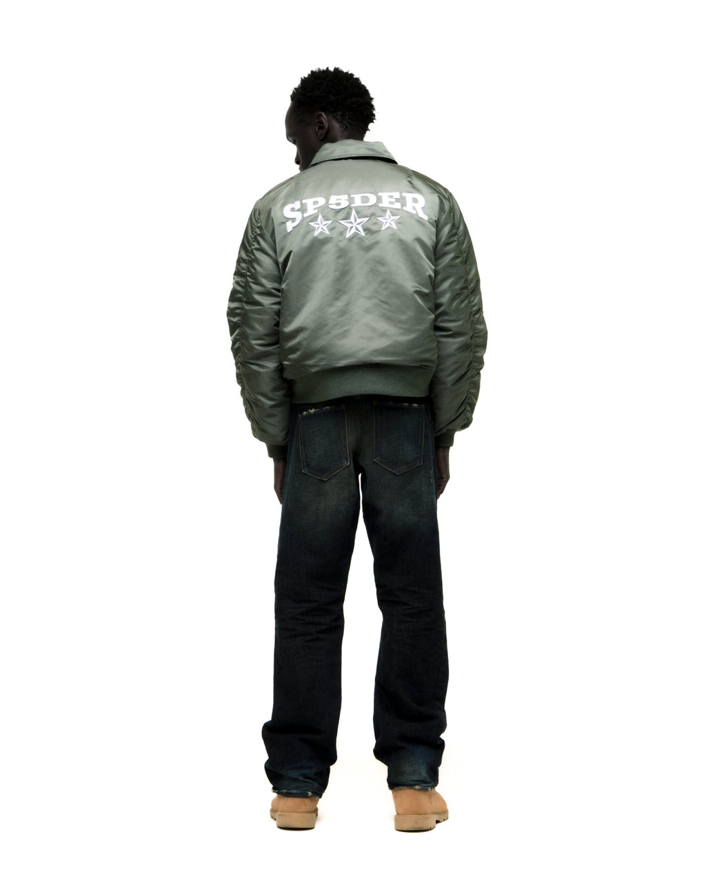 Man in green nylon bomber jacket with white SP5DER graphic and stars on back, dark jeans, and tan boots.