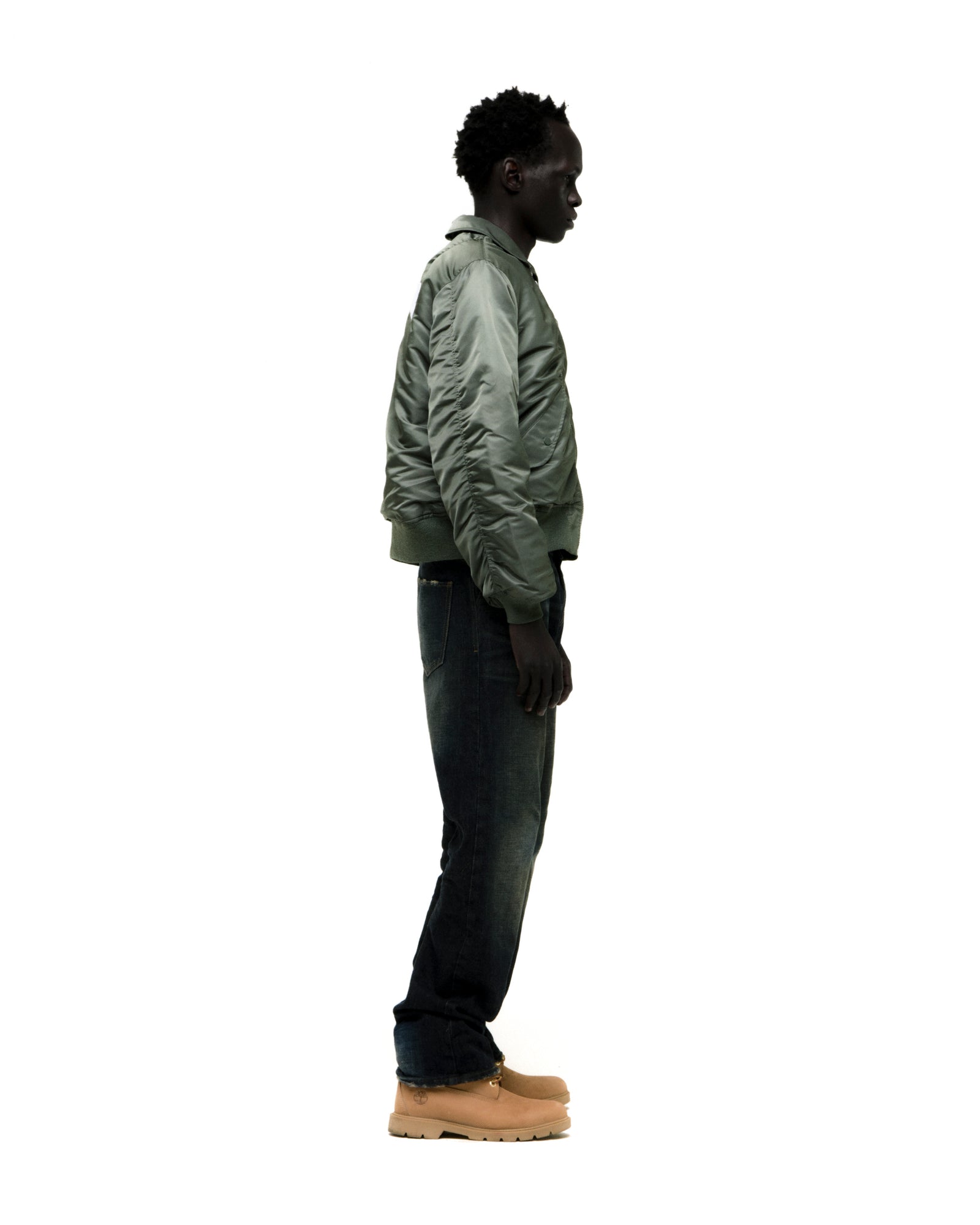 Man in a sage green nylon bomber jacket with ribbed cuffs, dark jeans, and tan boots.