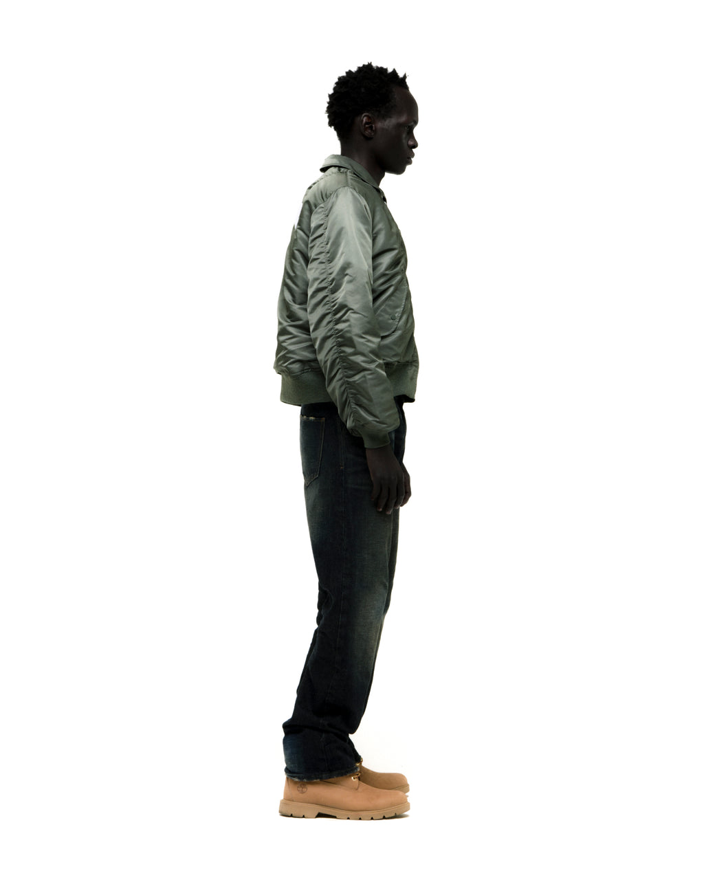 Man in a sage green nylon bomber jacket with ribbed cuffs, dark jeans, and tan boots.
