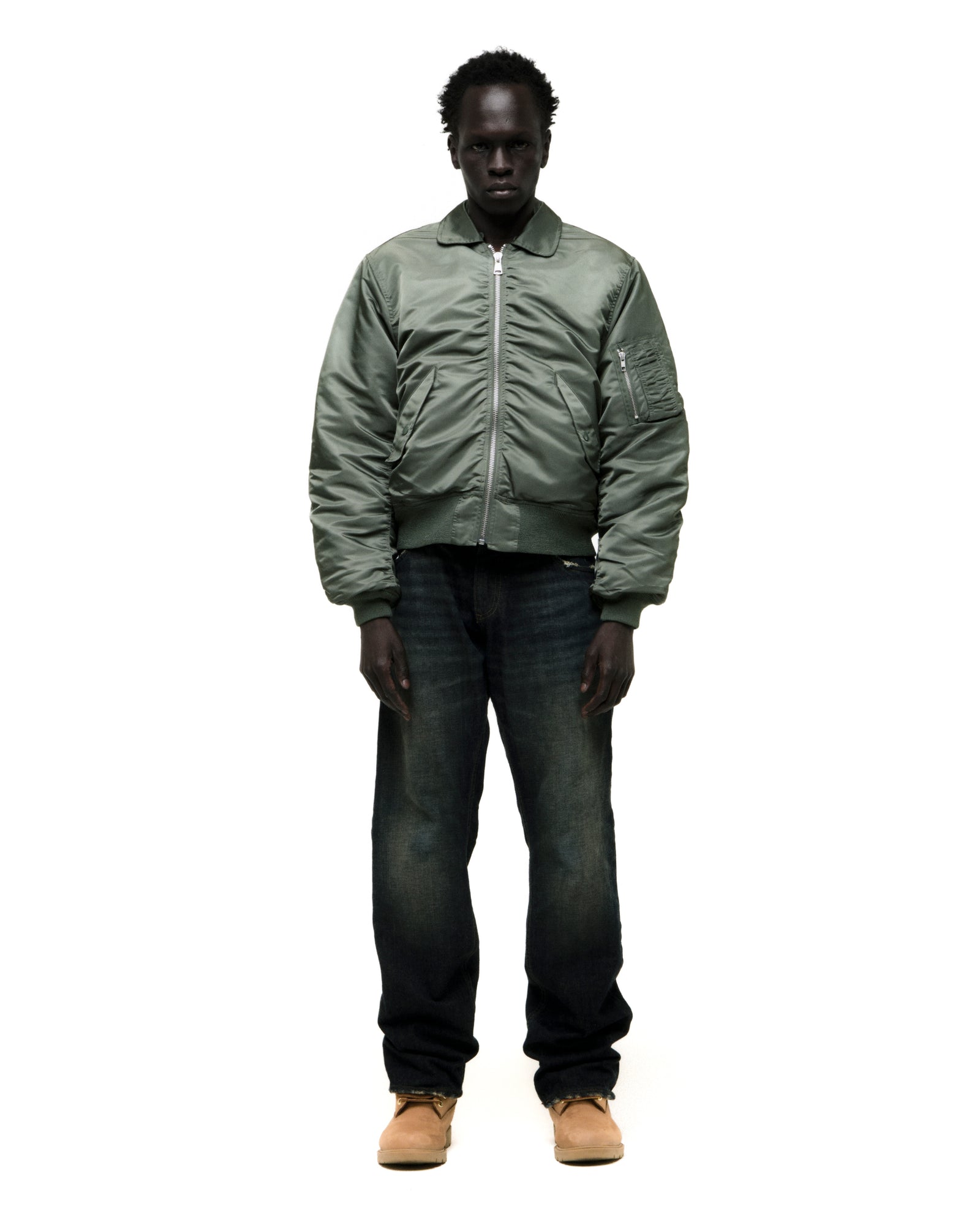 Man wearing a sage green nylon bomber jacket, dark jeans, and tan boots.