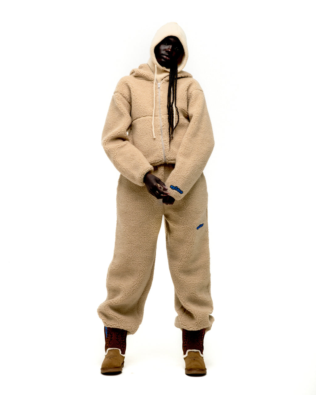 Beige teddy fleece zip-up hooded jacket and matching pants with blue logo patches, worn by a model.