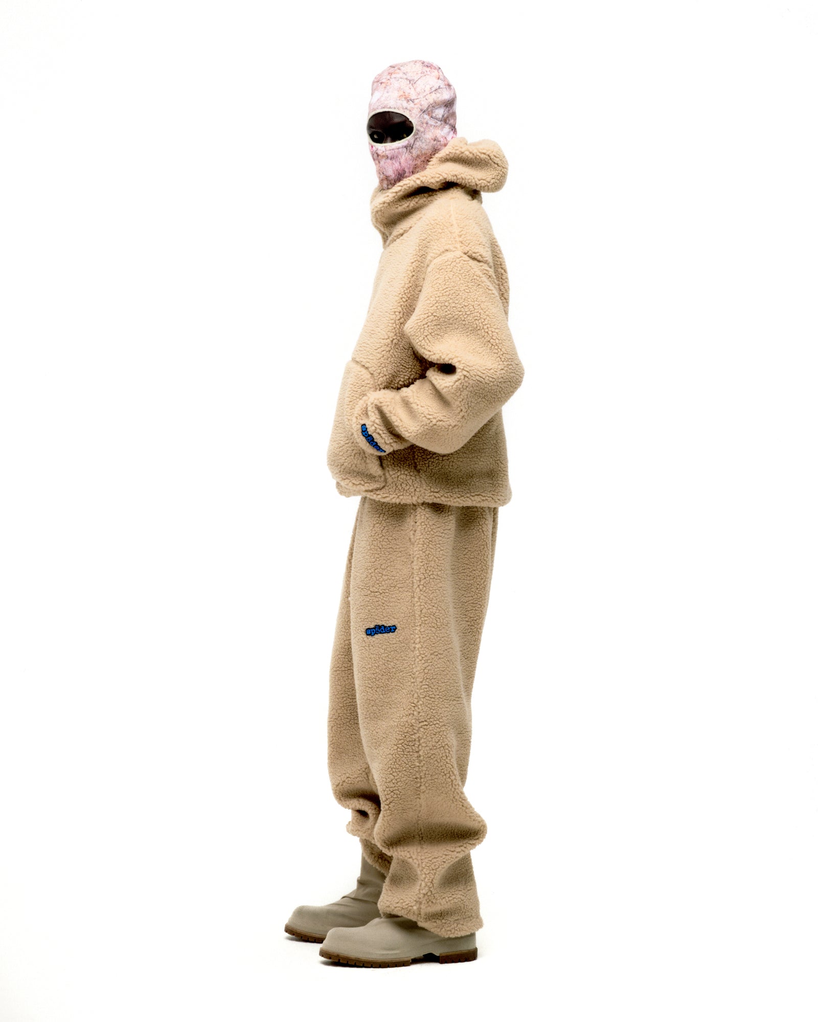 Beige teddy fleece zip-up jacket and matching pants with blue logo, worn by a person in a pink balaclava.