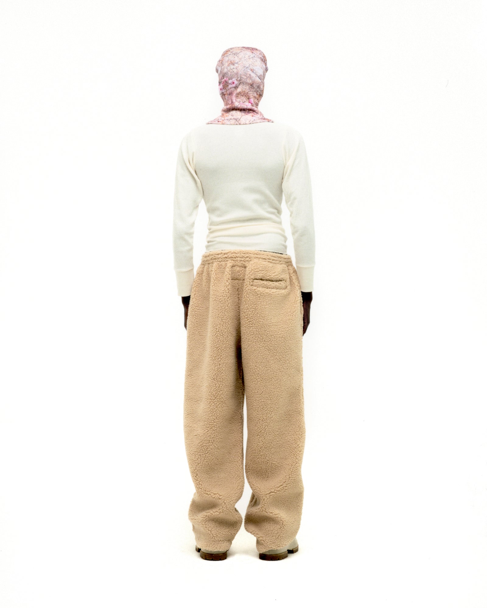 Rear view of a person wearing beige teddy fleece pants with a rubberized logo patch and a white long-sleeve shirt.