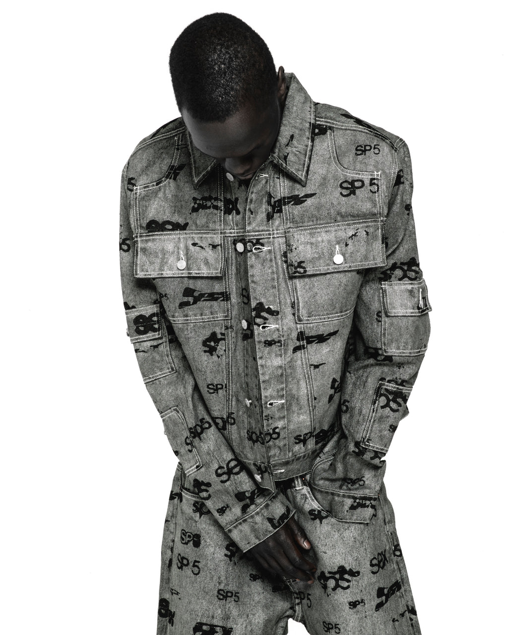 Gray denim trucker jacket with all-over black "SP5" print, silver buttons, and red contrast stitching.