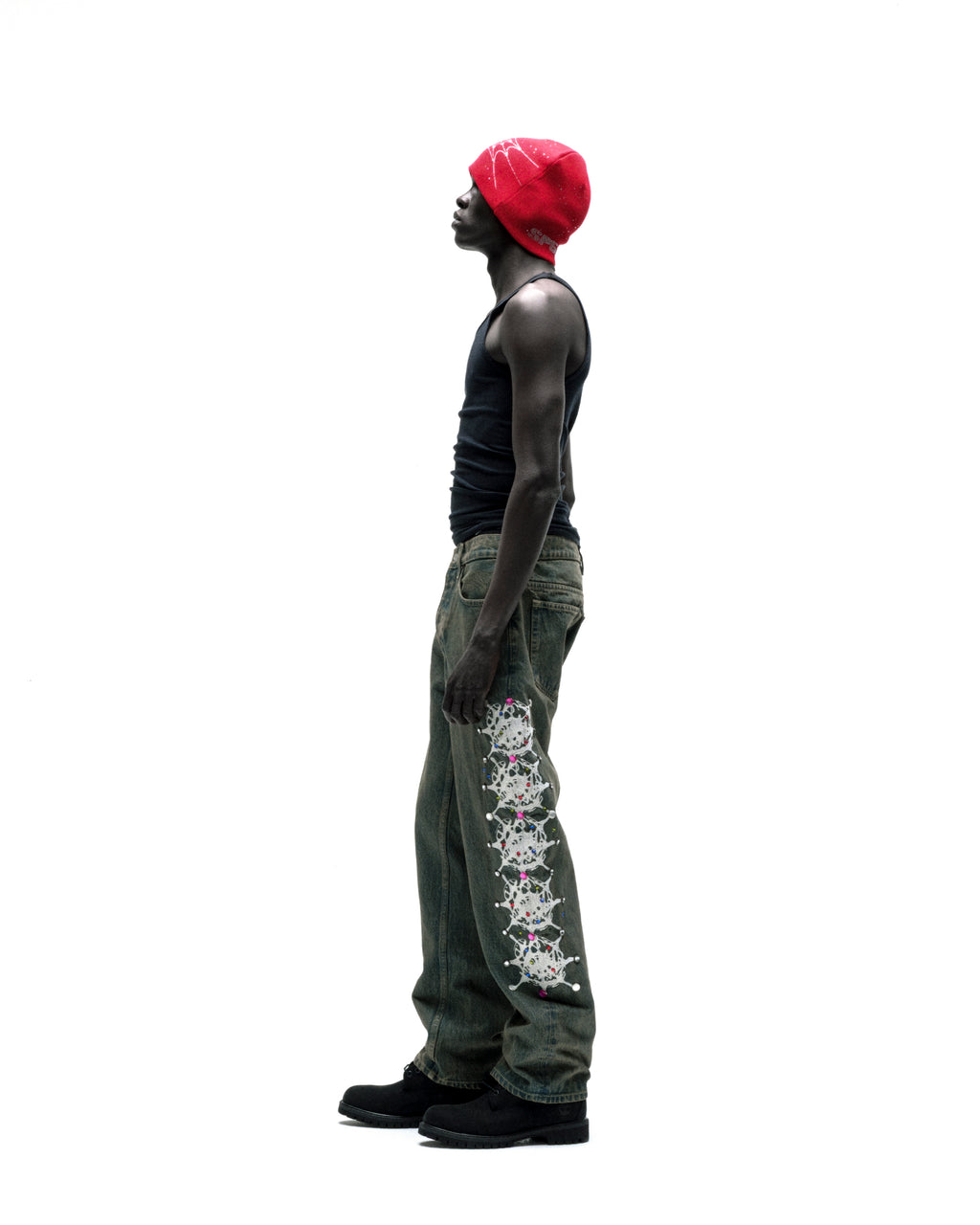 Man in vintage wash denim pants with web print and rhinestone embellishments, black tank top, and red beanie.