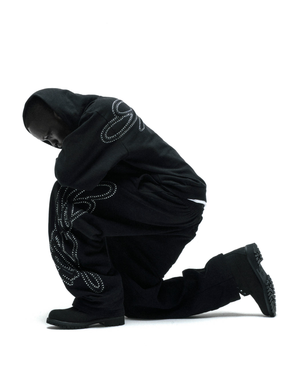 Black rhinestone script zip hoodie and sweatpants on a kneeling model.