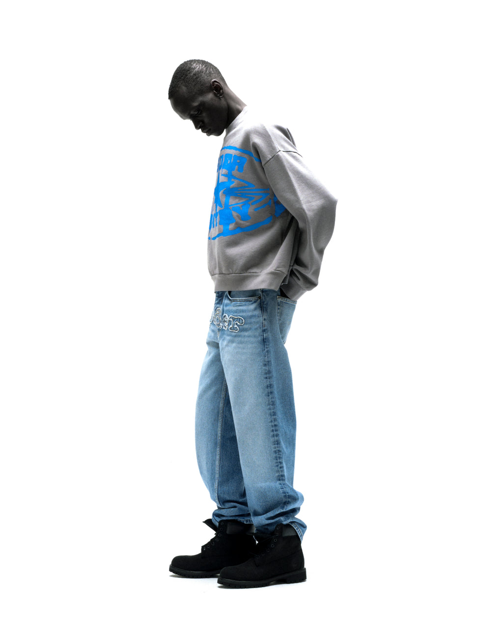 Man in grey SP5DER CUNTRY CREWNECK with blue graphic, light wash jeans, and black boots.