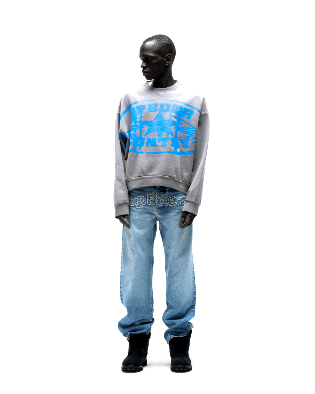 Gray SP5DER CUNTRY CREWNECK with blue graphic print, paired with light wash jeans and black boots.