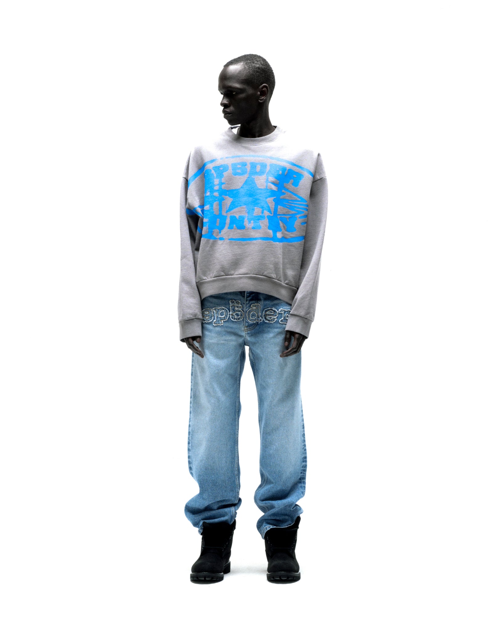 Gray SP5DER CUNTRY CREWNECK with blue graphic print, paired with light wash jeans and black boots.