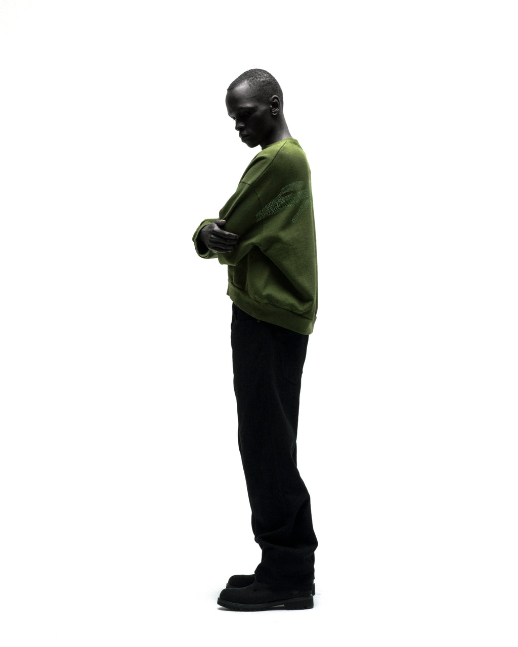 Model wearing an olive green RHINESTONE TM CREWNECK with rhinestone graphic, black pants, and shoes.
