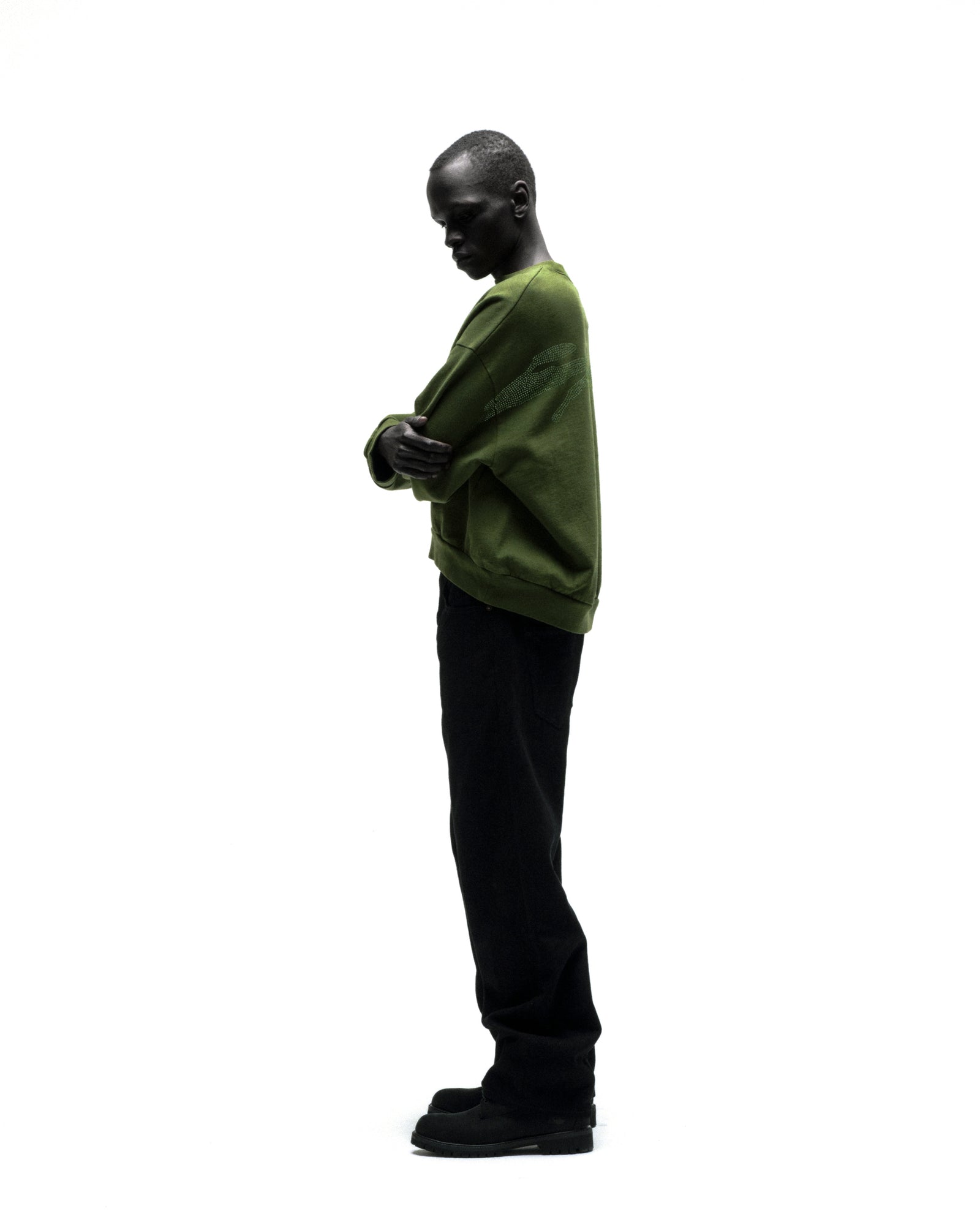 Model wearing an olive green RHINESTONE TM CREWNECK with rhinestone graphic, black pants, and shoes.