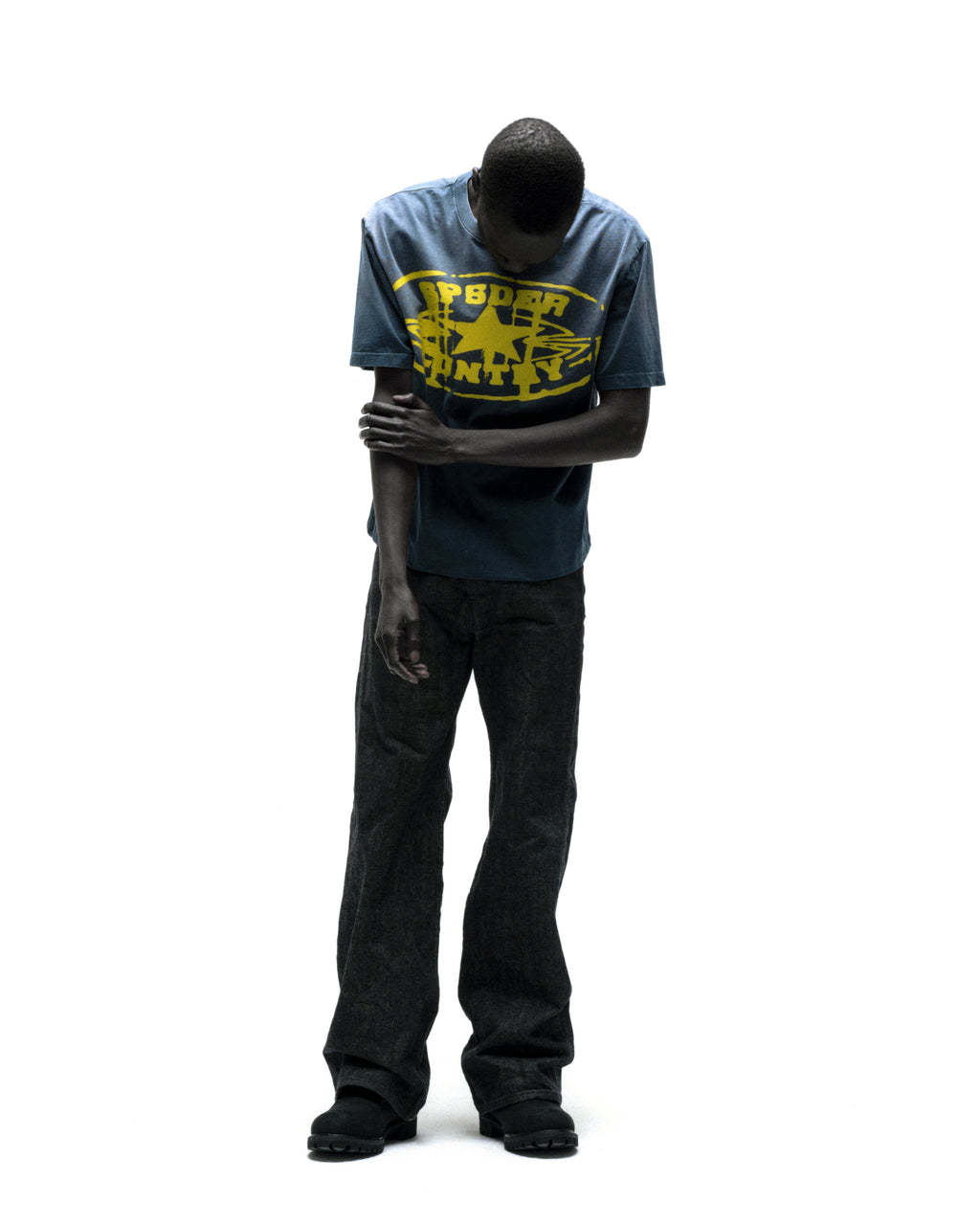 Man in faded blue cotton SP5DER CUNTRY TEE with yellow graphic print and dark jeans.