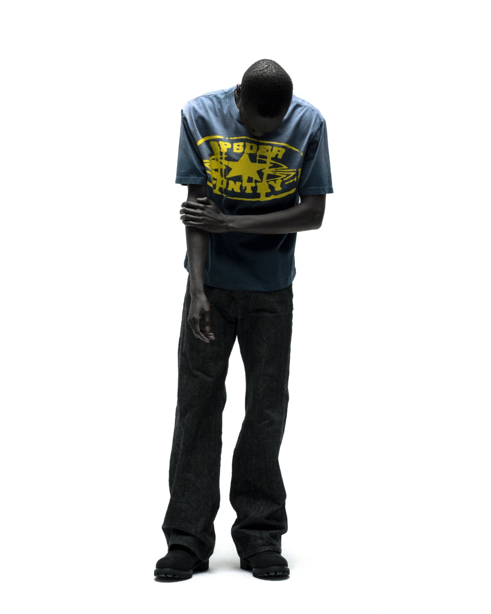 Man in faded blue cotton SP5DER CUNTRY TEE with yellow graphic print and dark jeans.