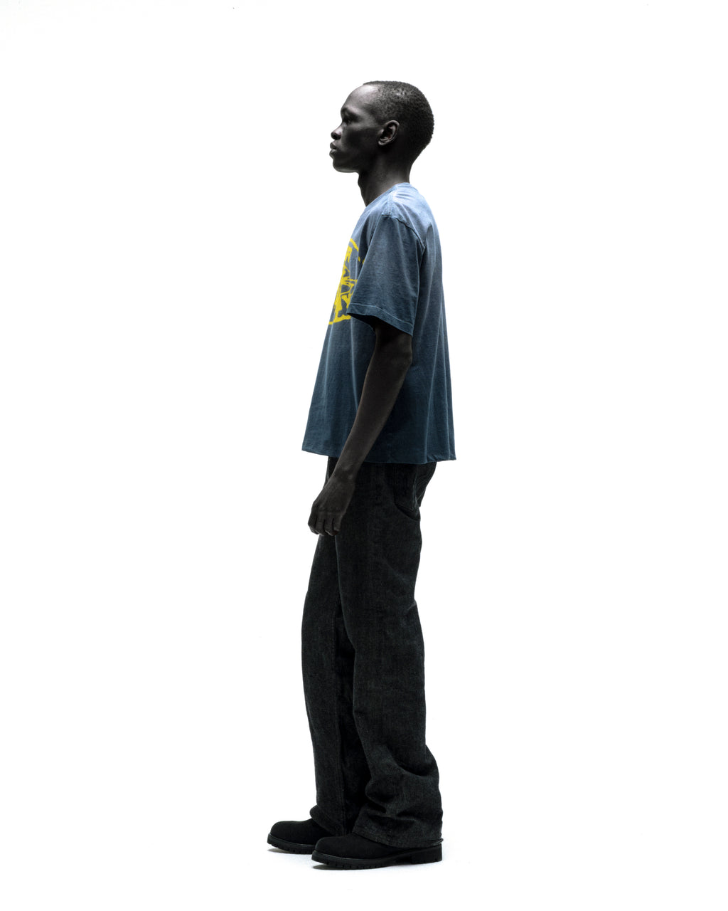 Man in faded blue SP5DER CUNTRY TEE with yellow graphic, dark jeans, and black shoes.