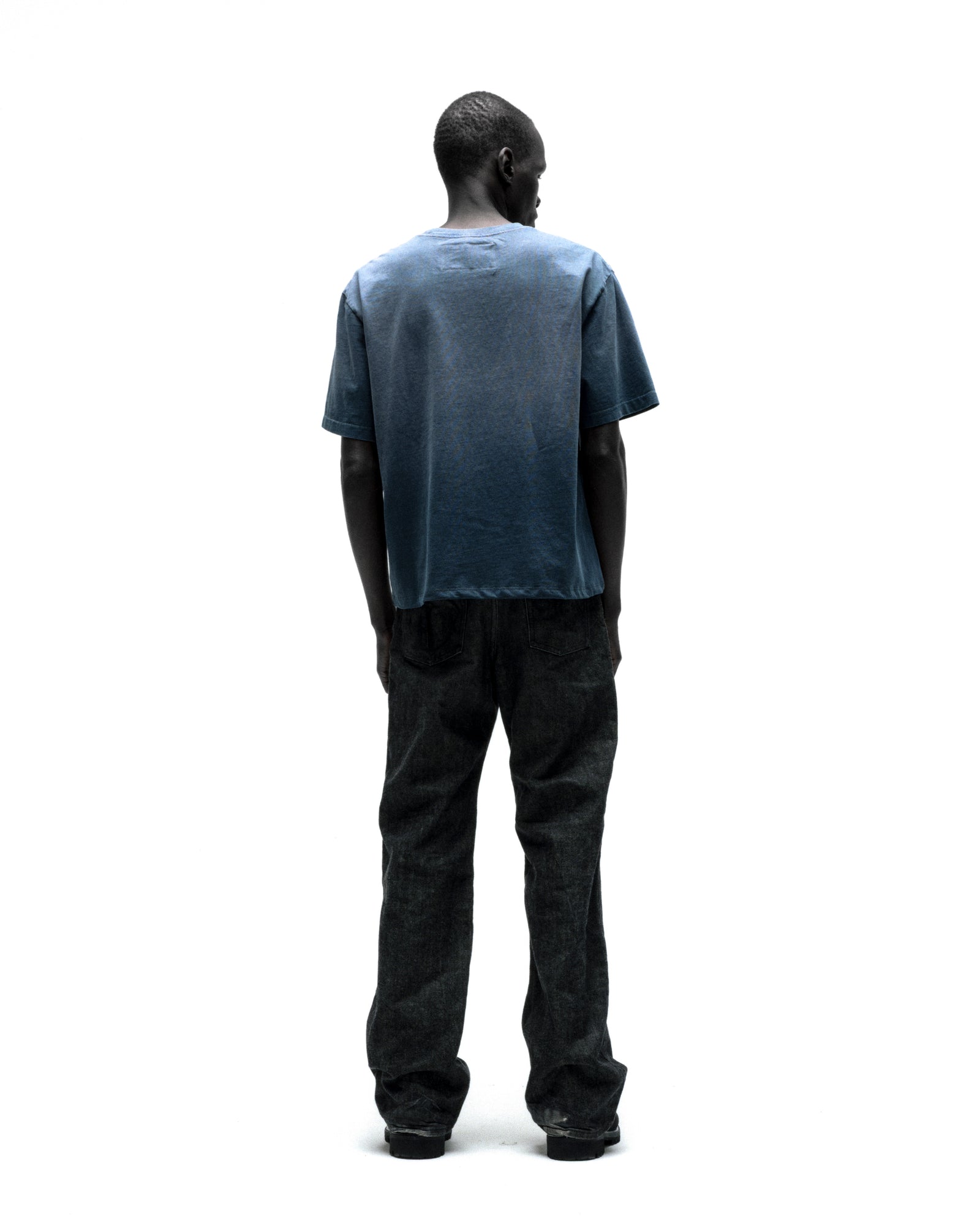 Man from behind wearing a faded blue cotton SP5DER CUNTRY TEE with a graphic print and dark jeans.