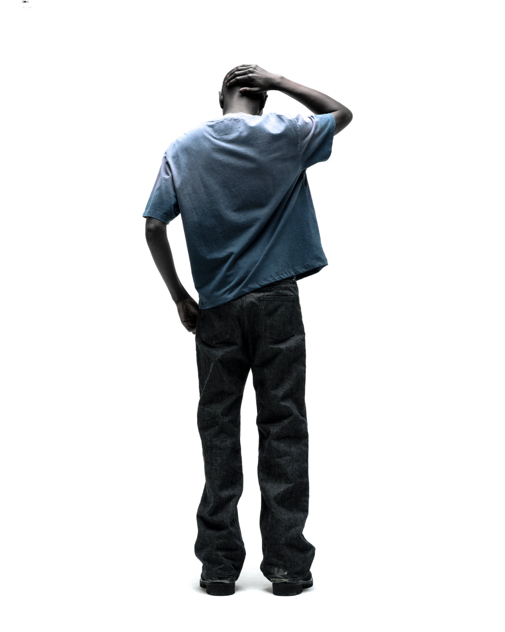 Man in faded blue FORM 5 TEE and dark jeans, back to viewer, hand on head.