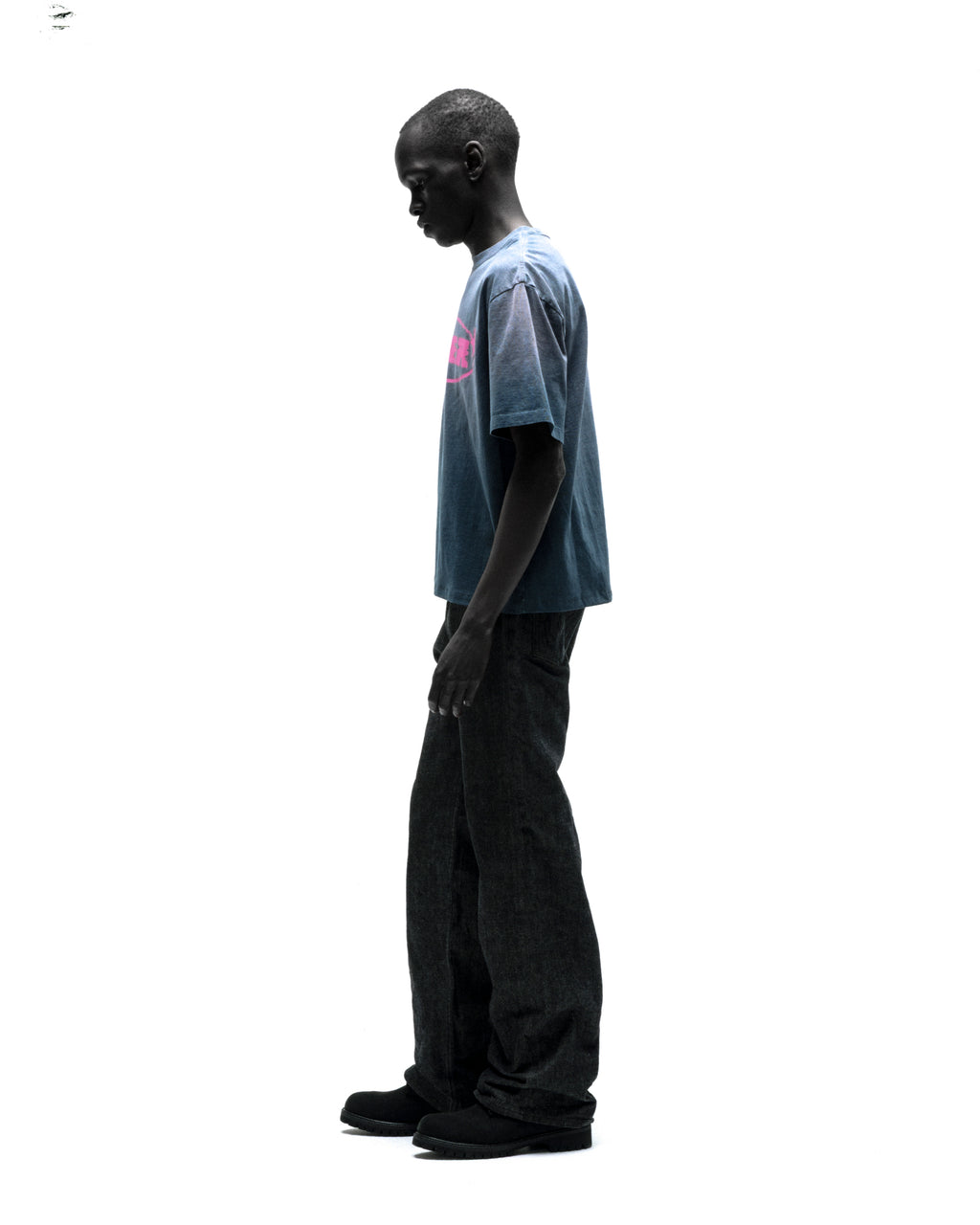 Man in faded blue FORM 5 TEE with pink graphic, black jeans, and shoes, side profile.