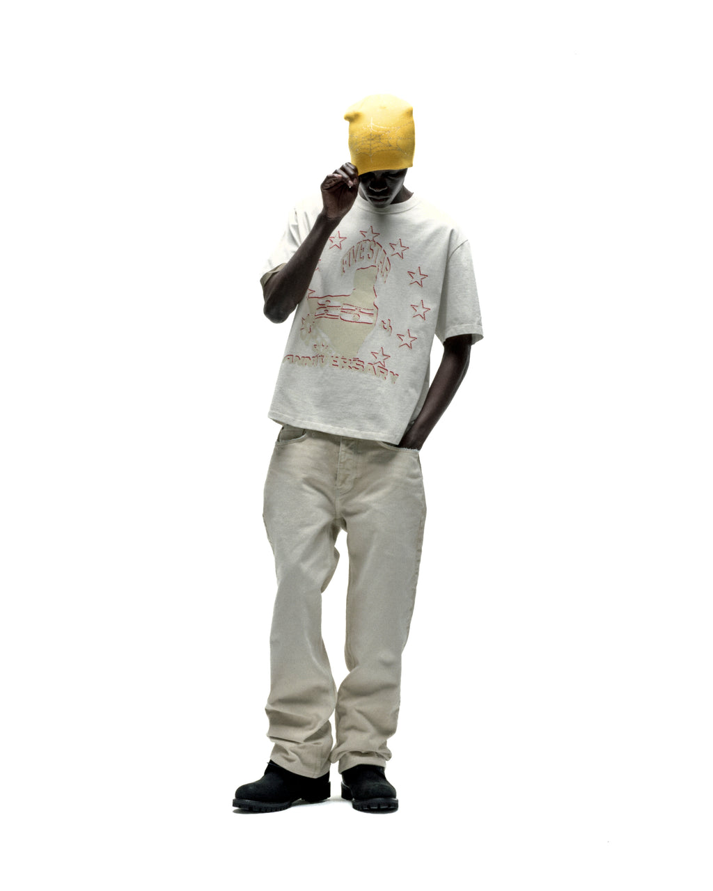 Man in a cream cotton ANNIVERSARY TEE with red graphic print, light beige jeans, and a yellow beanie.