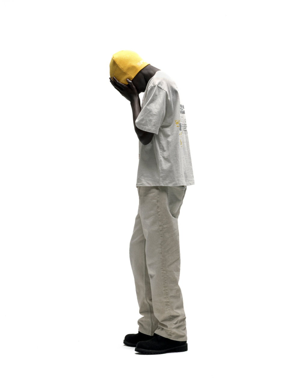 Man in a light grey cotton ANNIVERSARY TEE with graphic print, beige pants, and yellow hat.