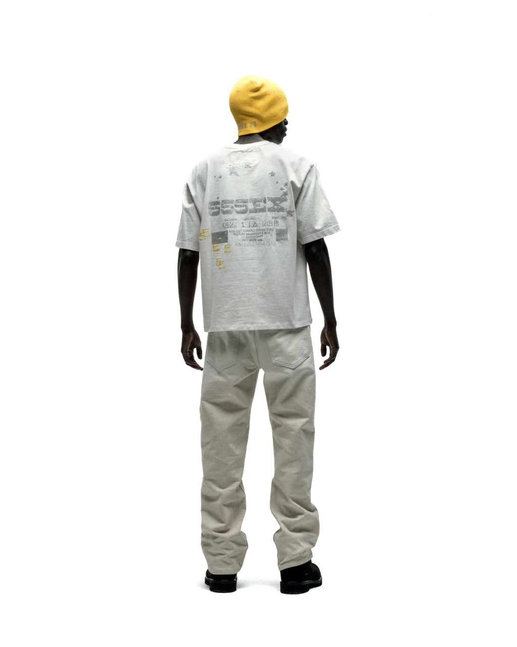 Man in a light grey cotton ANNIVERSARY TEE with back graphic print, light pants, and yellow beanie.