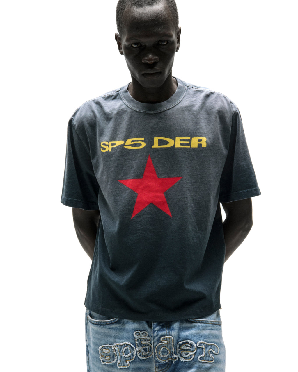 Dark-skinned man wearing a faded wash grey cotton tee with "SP5 DER" in yellow and a red star graphic.