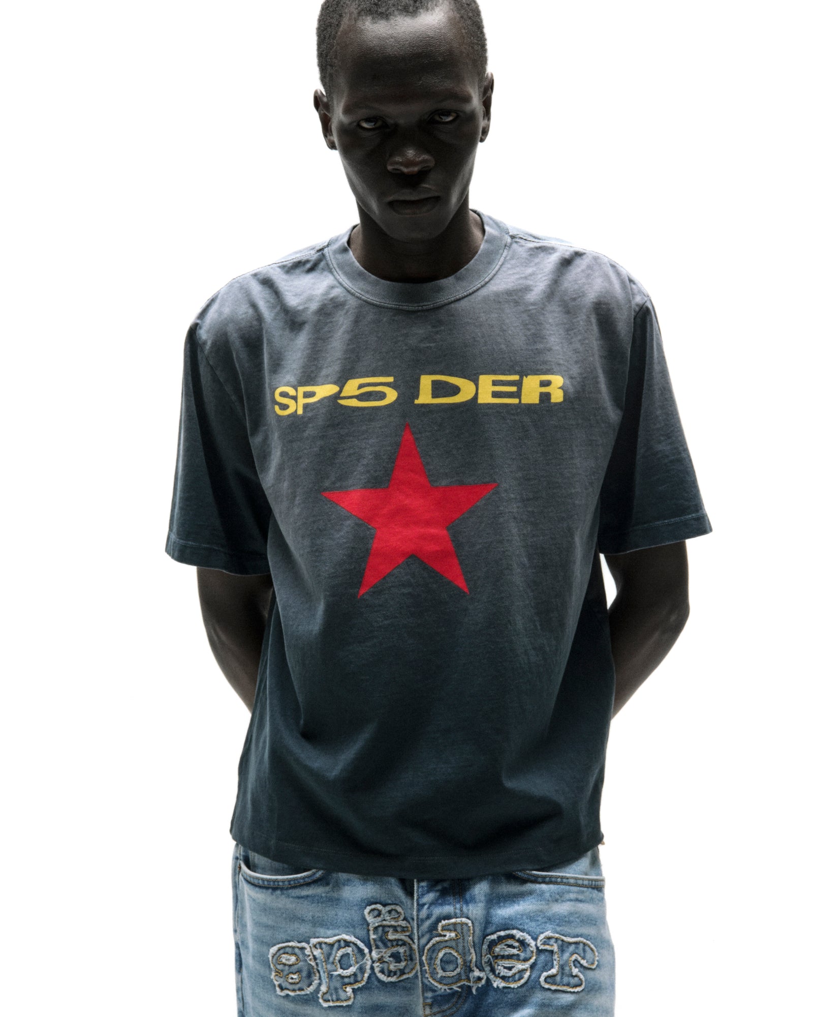 Dark-skinned man wearing a faded wash grey cotton tee with "SP5 DER" in yellow and a red star graphic.