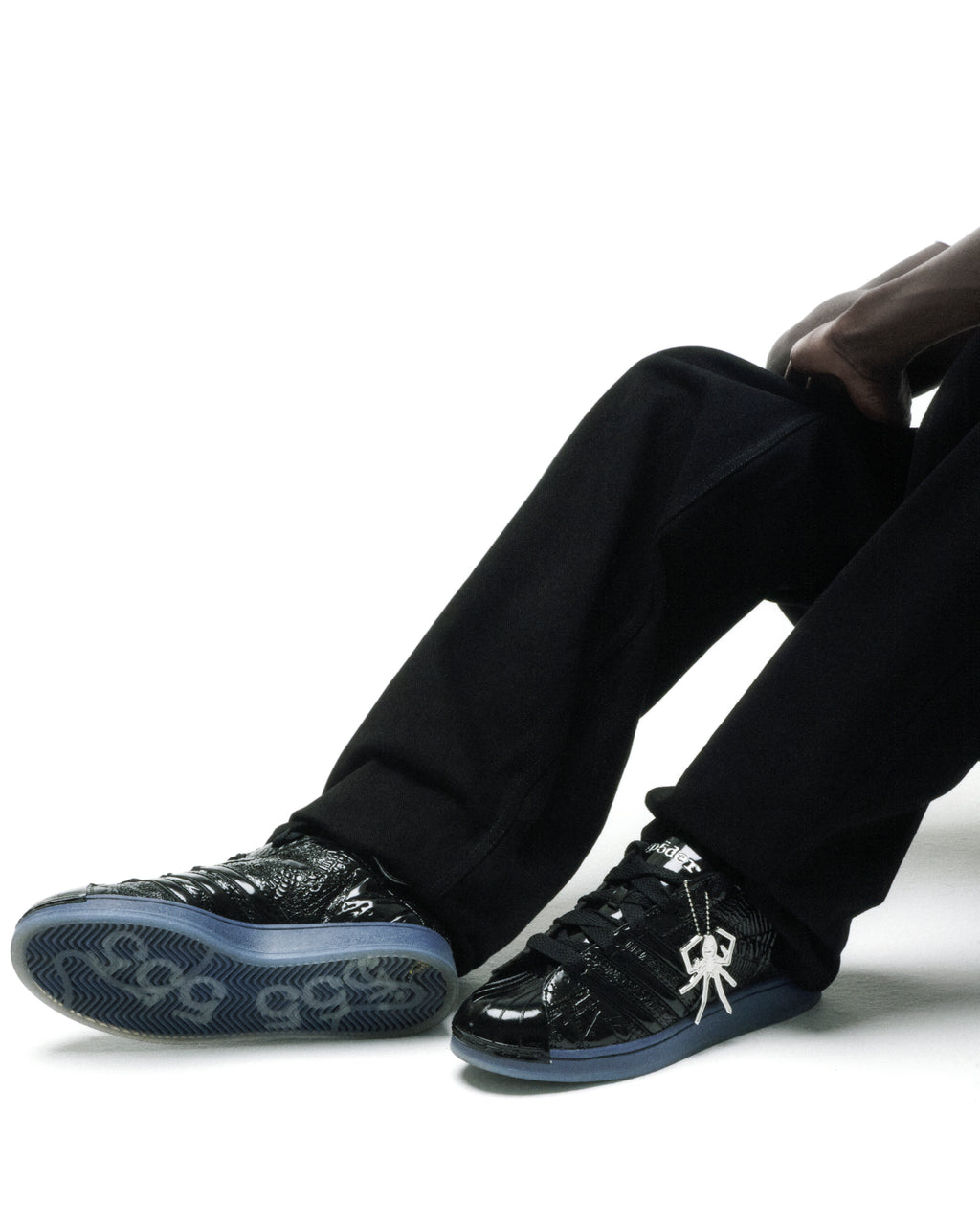 SP5DER x ADIDAS SUPERSTAR black glossy patent leather sneakers with blue soles and a silver spider charm.