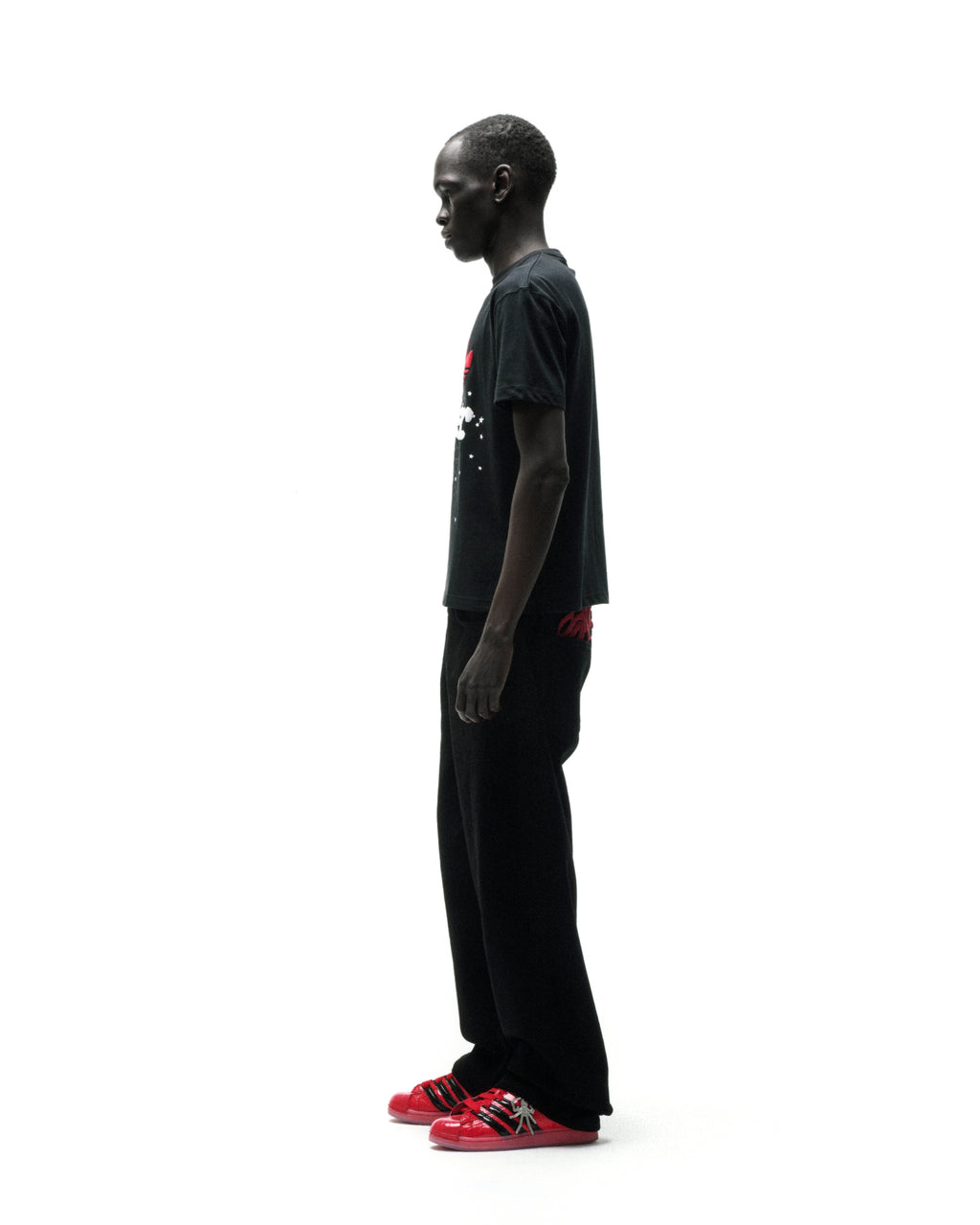 Man in black Adidas Souvenir Tee with Trefoil x SP5DER graphic, black pants, and red sneakers.