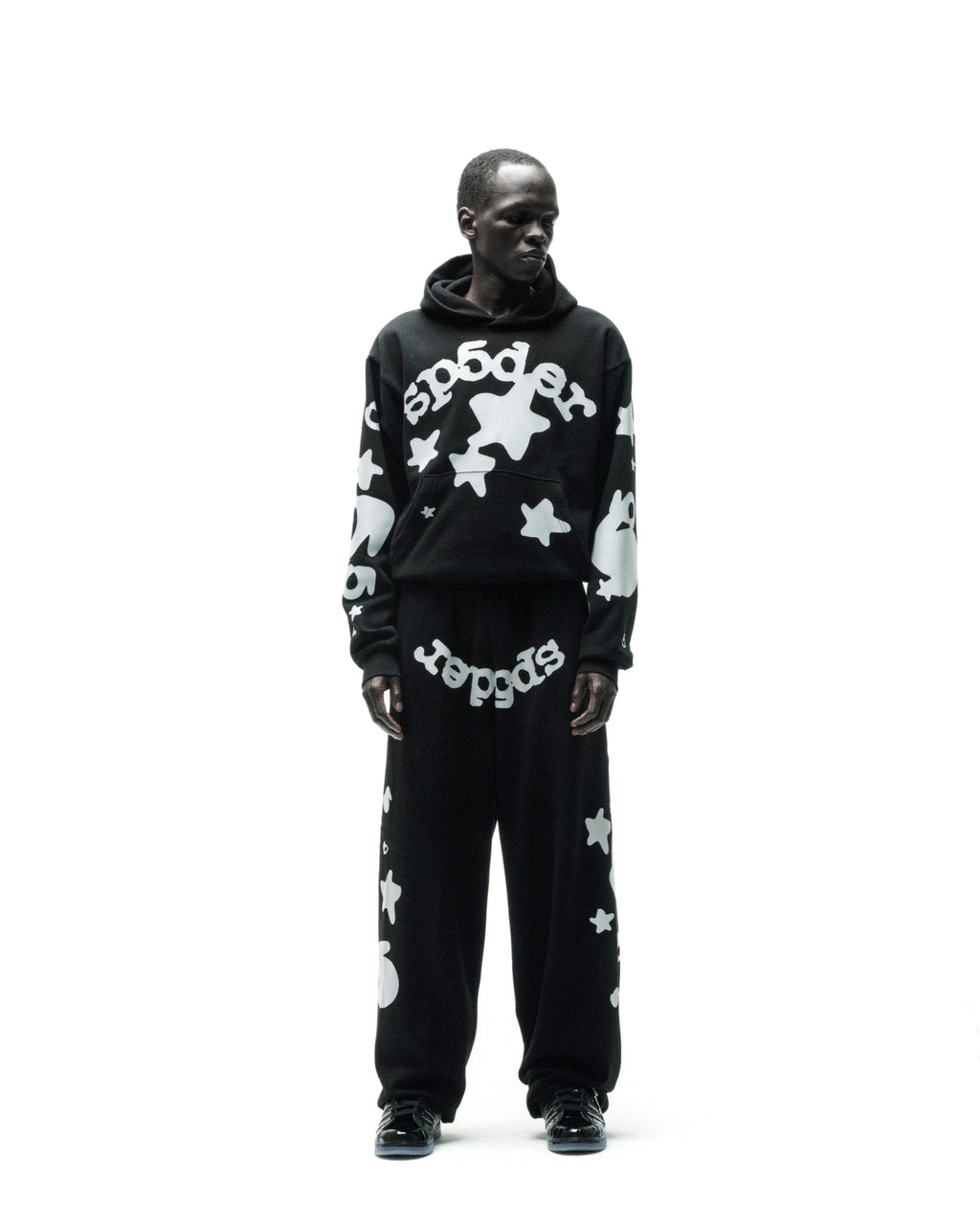 Man in black REFLECTIVE BELUGA HOODIE and sweatpants with white 'SP5DER' and star print.