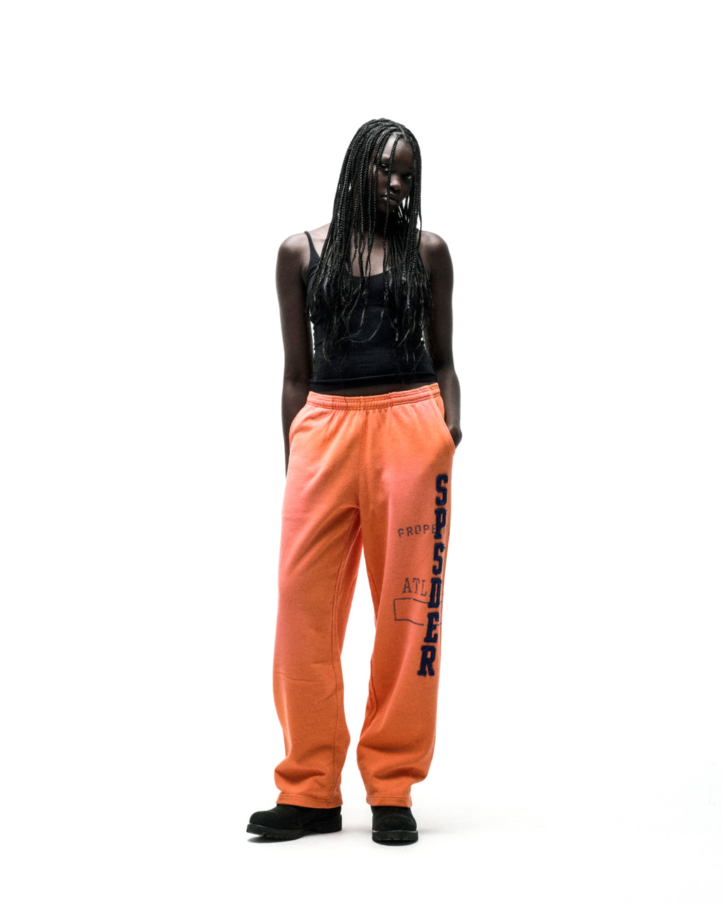 Model in orange wide-leg cotton fleece sweatpants with faded graphic print and raised chenille-style "SPSDER" lettering.